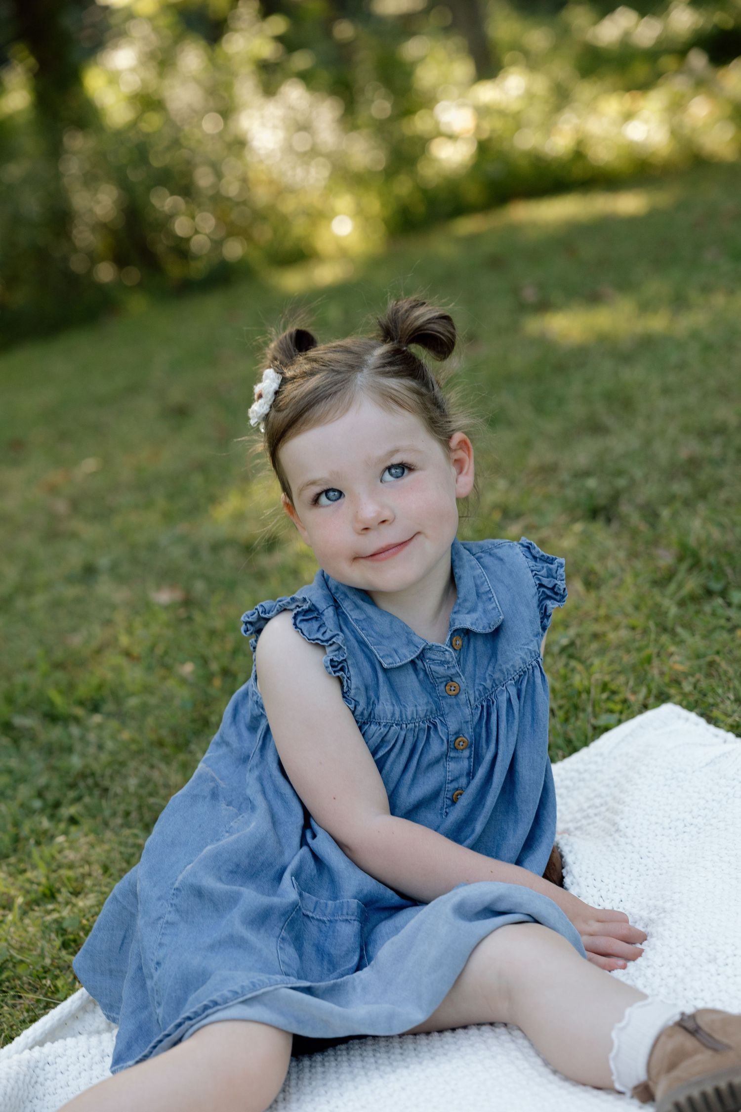 Adorable young girl in a blue denim dress sitting on a white blanket outdoors in a green park.