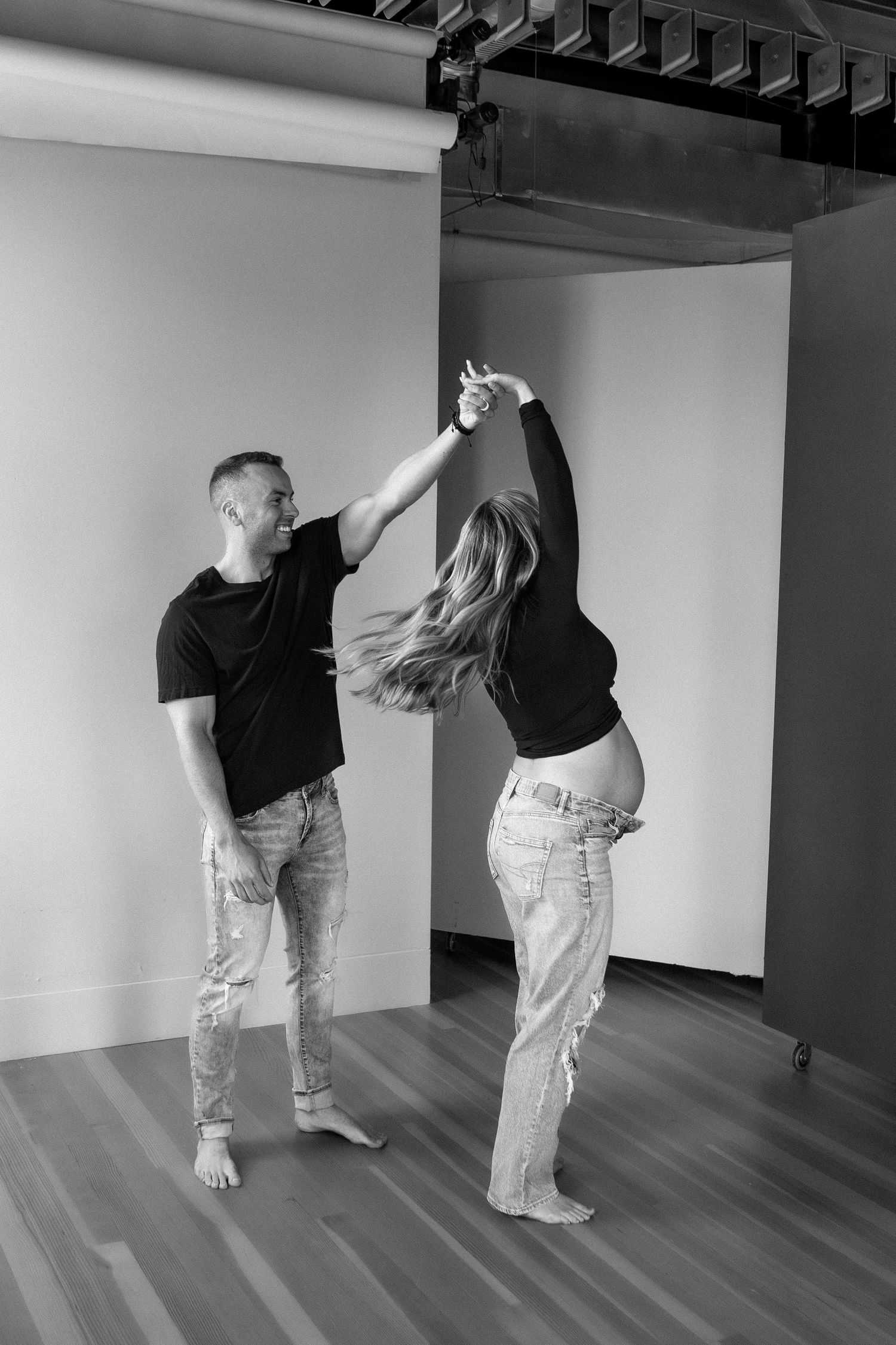 Black and white photo of a couple dancing together, with pregnant woman being twirled by her partner in a studio.