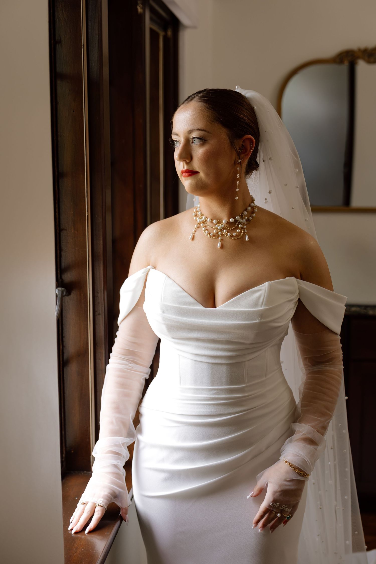 Elegant bride in off-shoulder white gown with gold jewelry and veil gazing out a window.