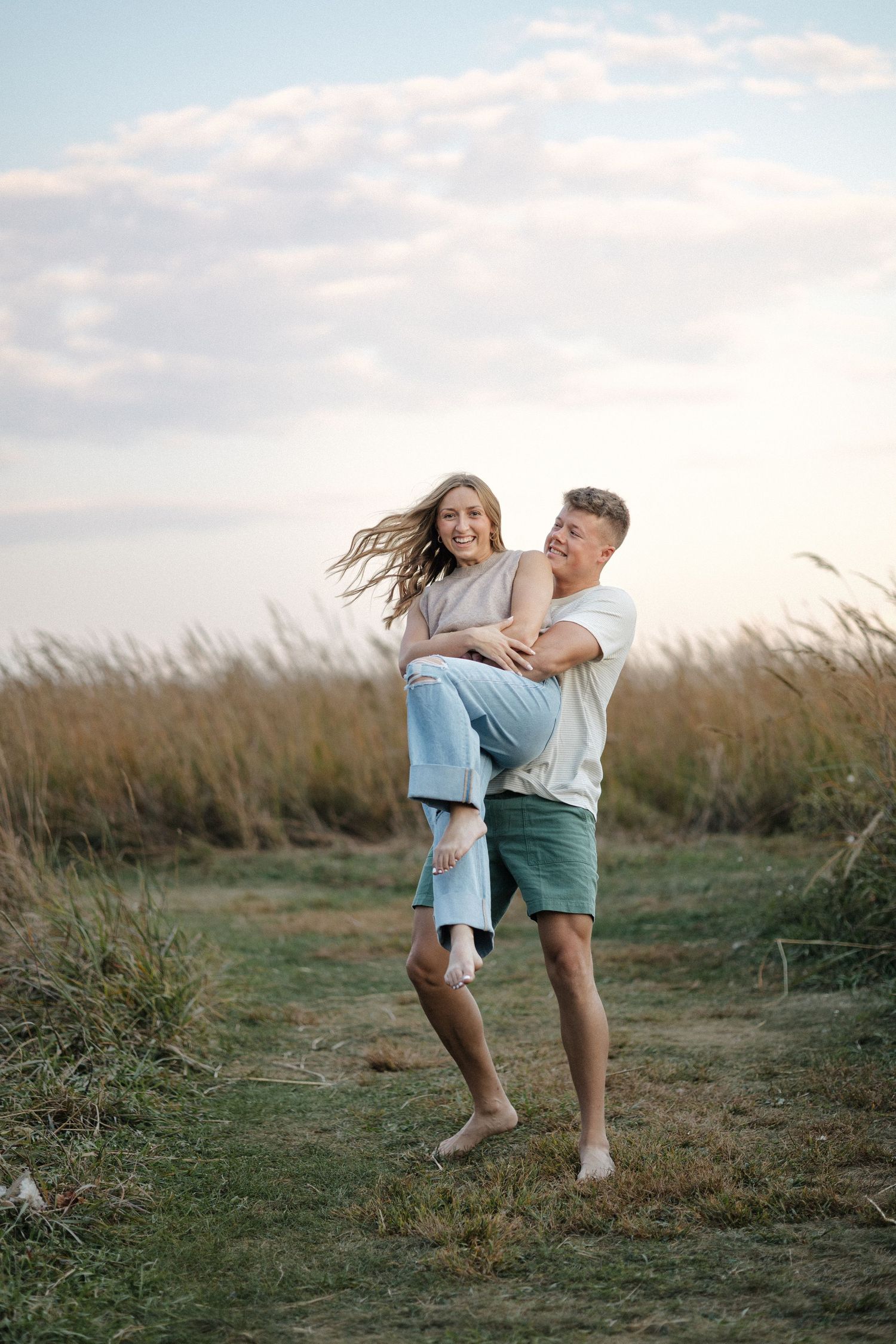 Man lifting smiling woman in his arms in golden field at dusk, both barefoot.