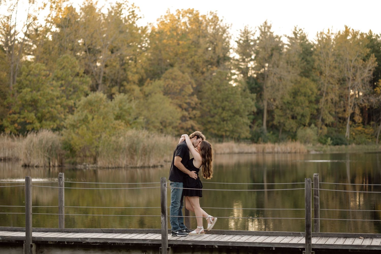 Couple sharing a romantic kiss on a wooden dock overlooking a serene lake surrounded by autumn foliage.