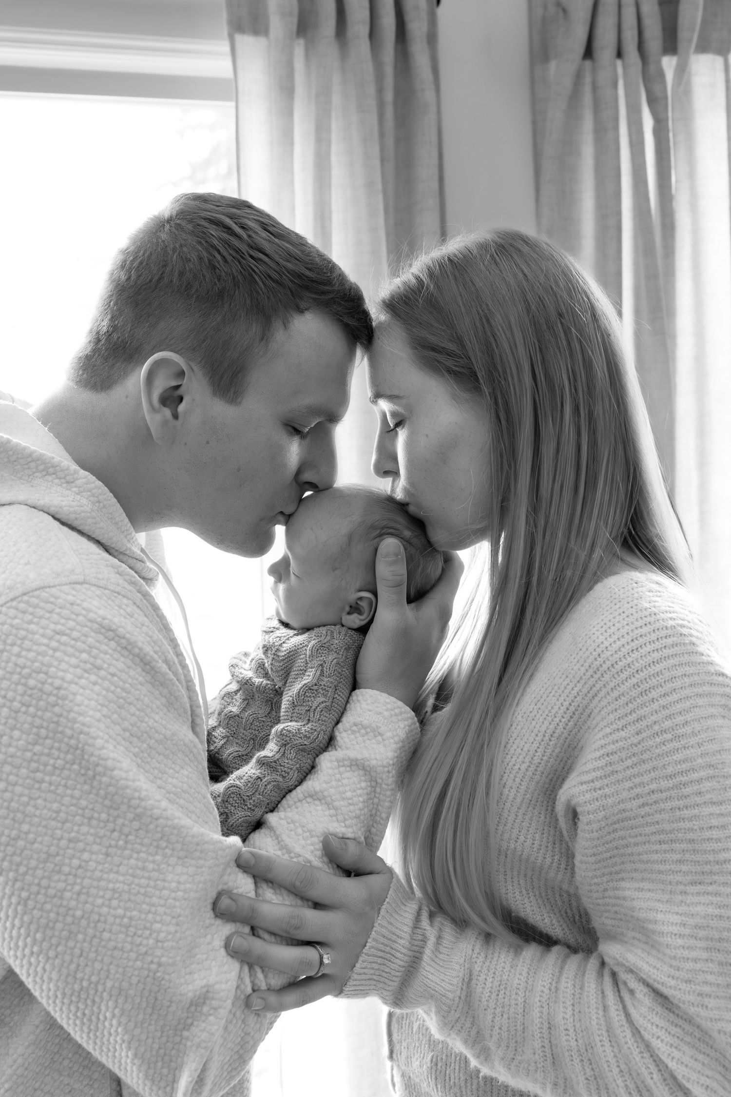 Black and white photo of new parents kissing over their bundled newborn baby in a softly lit room.