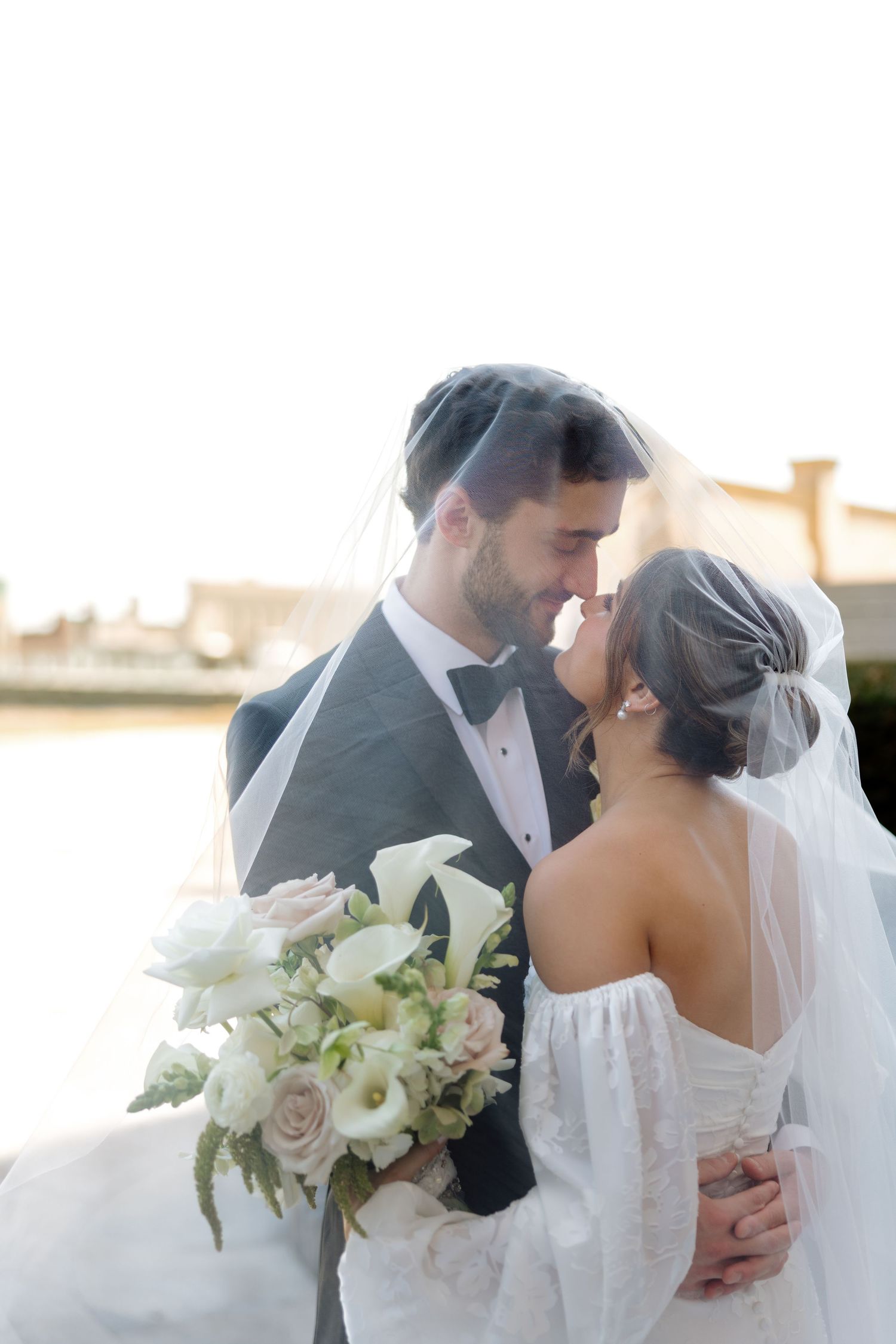 Bride and groom nearly kiss under veil while bride holds white calla lily bouquet outdoors.