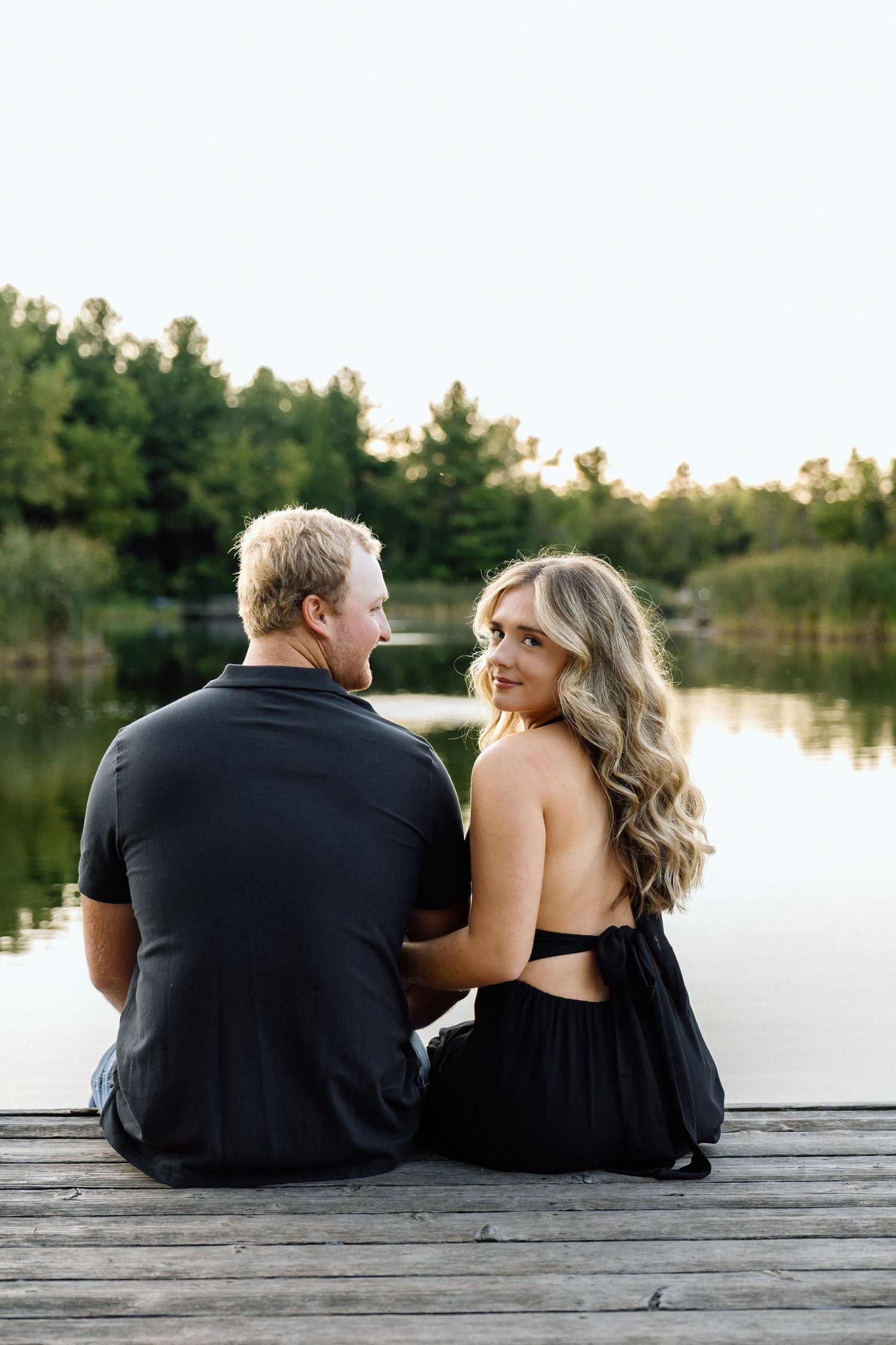 Couple in black outfits sitting on a wooden dock by a calm lake, woman turning to smile at camera.