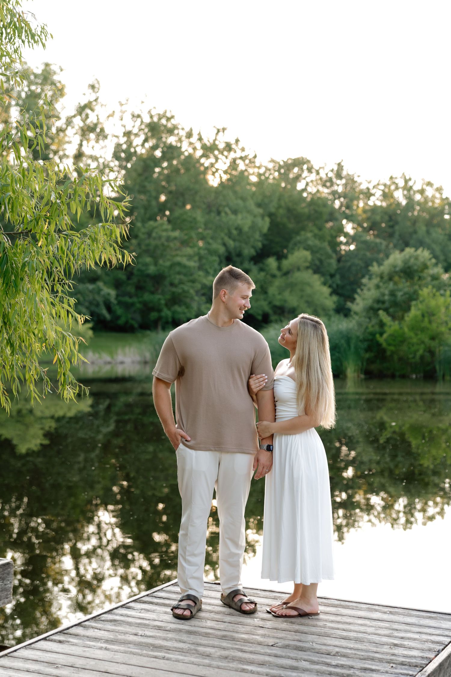 Couple standing on a wooden dock by a calm lake, surrounded by lush green trees, sharing a romantic moment.