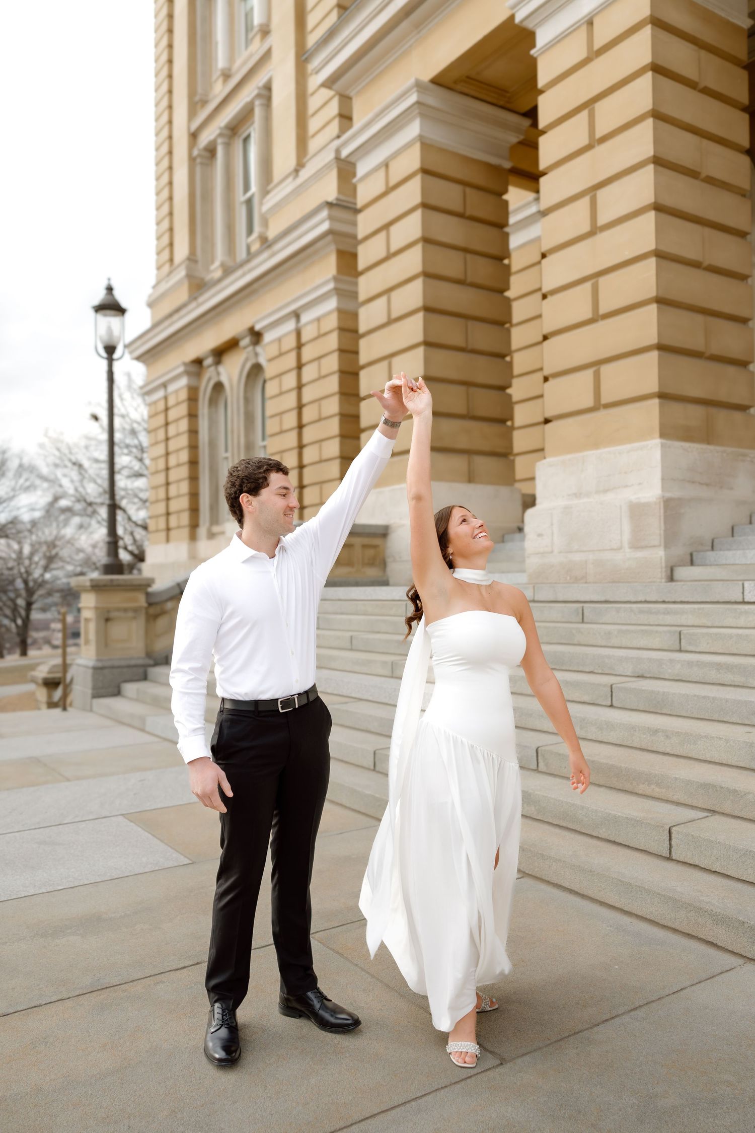 Bride in white gown and groom in white shirt dance joyfully outside a classic stone building.