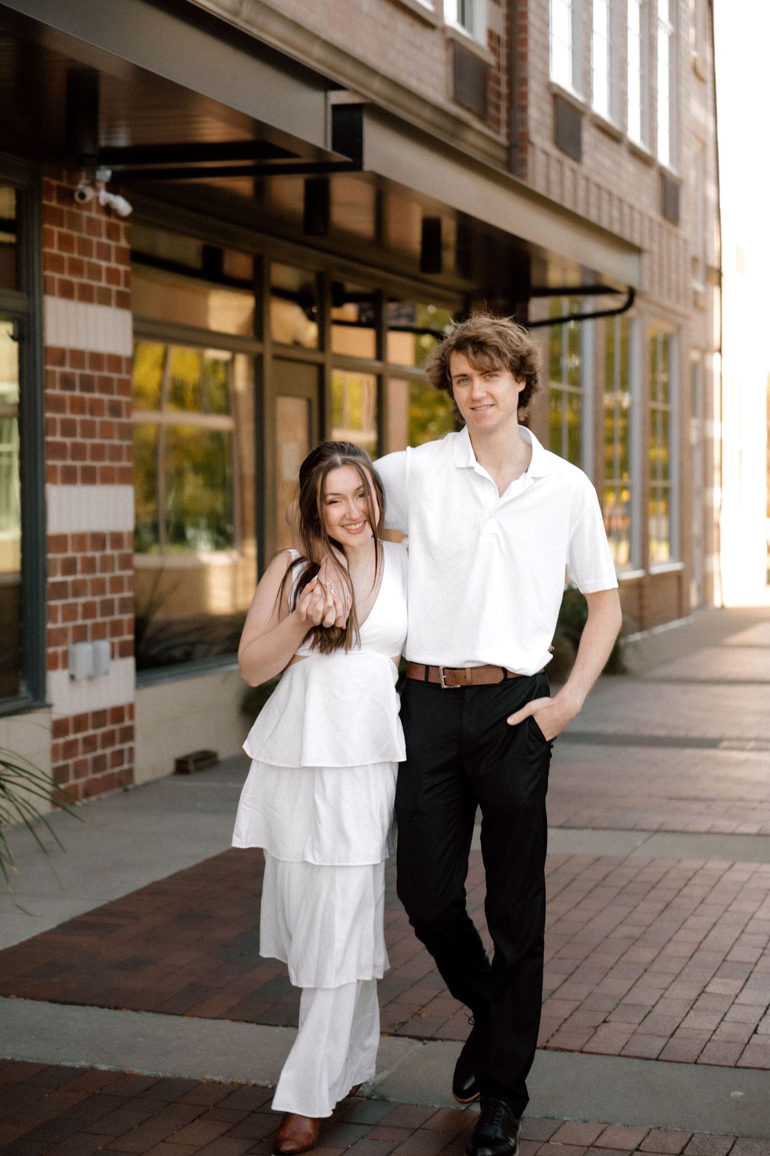 Young couple in white outfits strolling along brick sidewalk beside historic building.