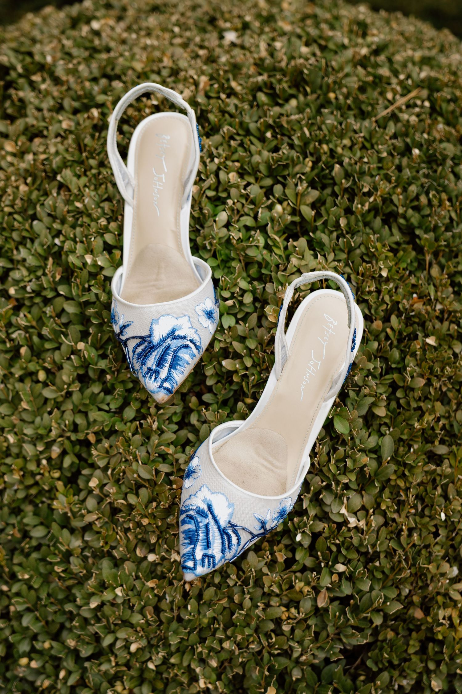 Elegant white and blue floral embroidered slingback wedding shoes displayed on lush green boxwood hedge.
