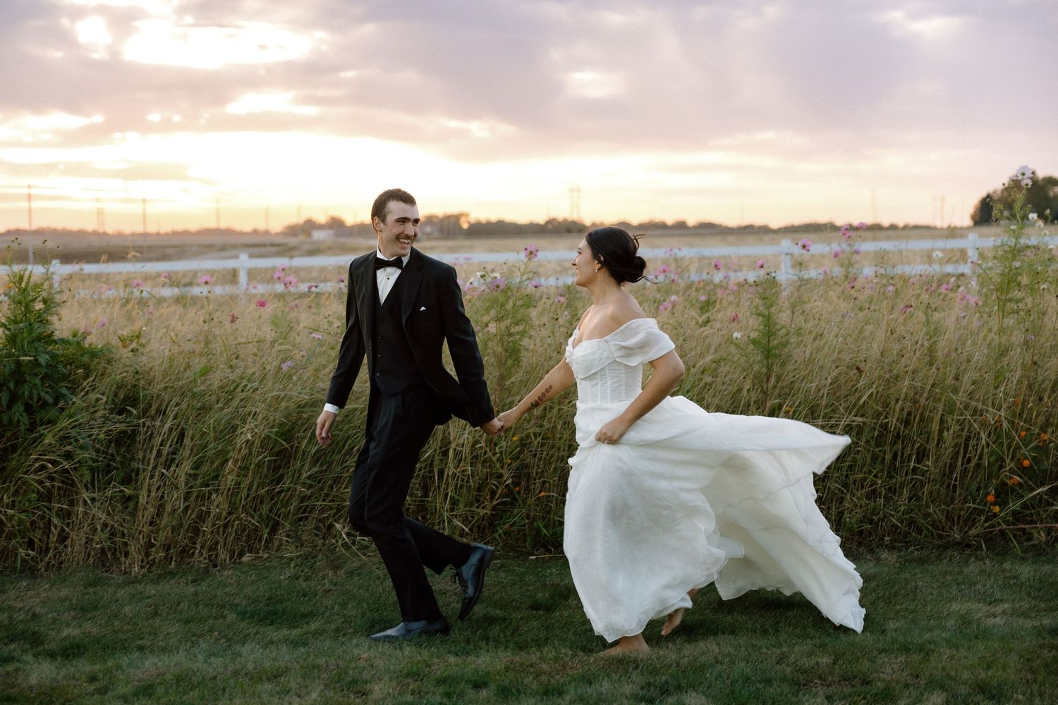 Bride and groom hold hands running through tall grass at sunset near a white fence.