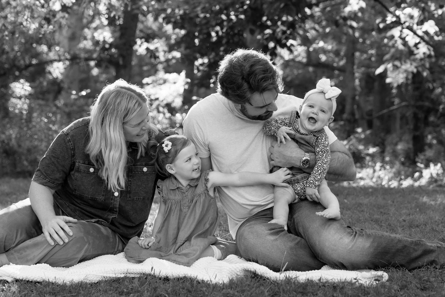 Black and white candid family photo of parents laughing with two young daughters on blanket.