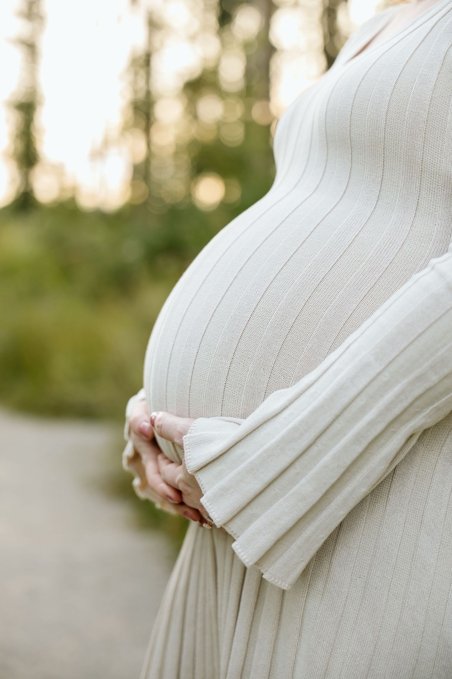 Close-up of pregnant woman in white ribbed knit dress gently cradling baby bump outdoors in soft light.