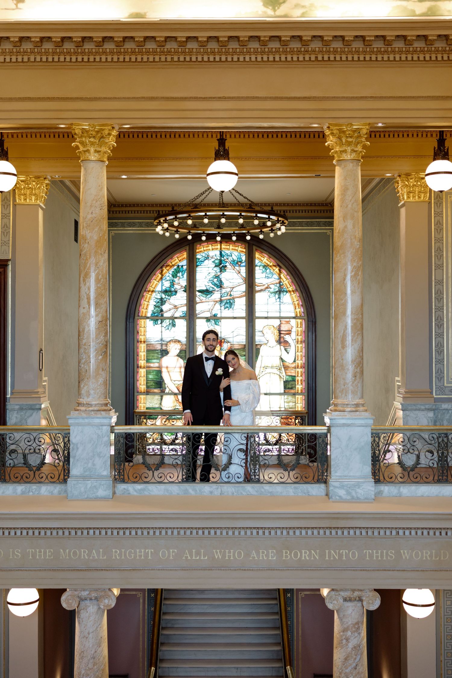Bride and groom posing on a balcony with stained glass window in a grand historic venue.