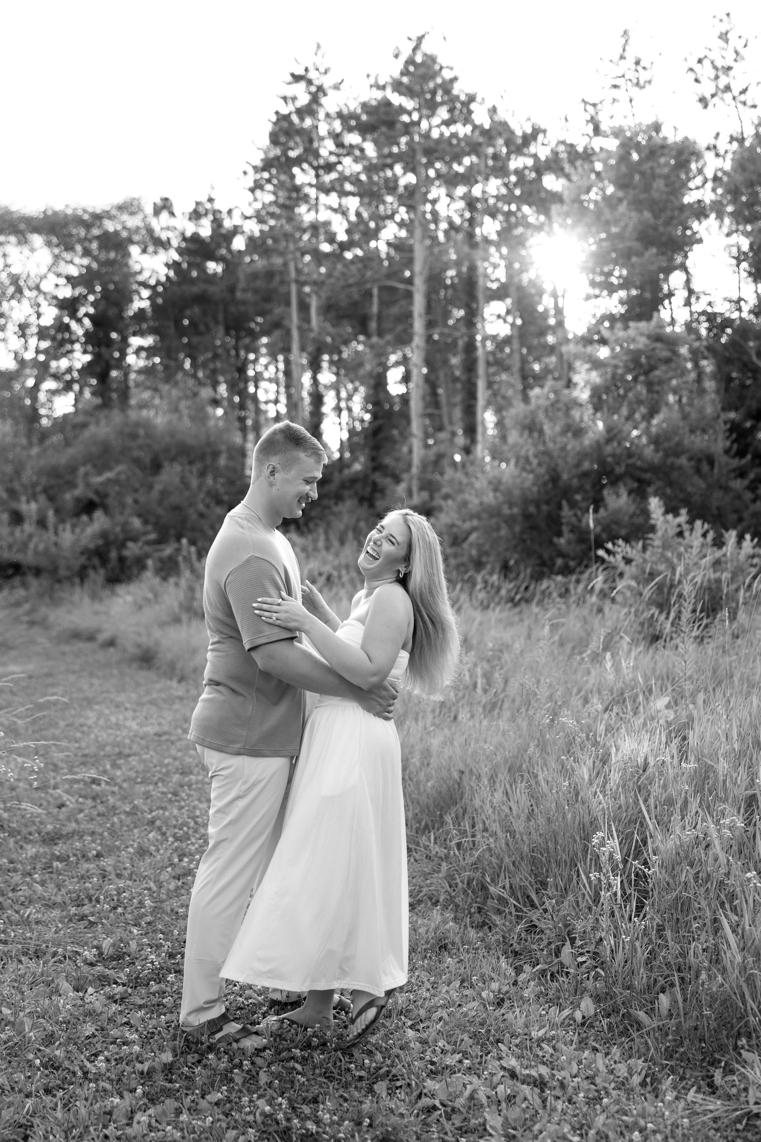 Black and white photo of a couple embracing and laughing in a sunlit forest meadow.