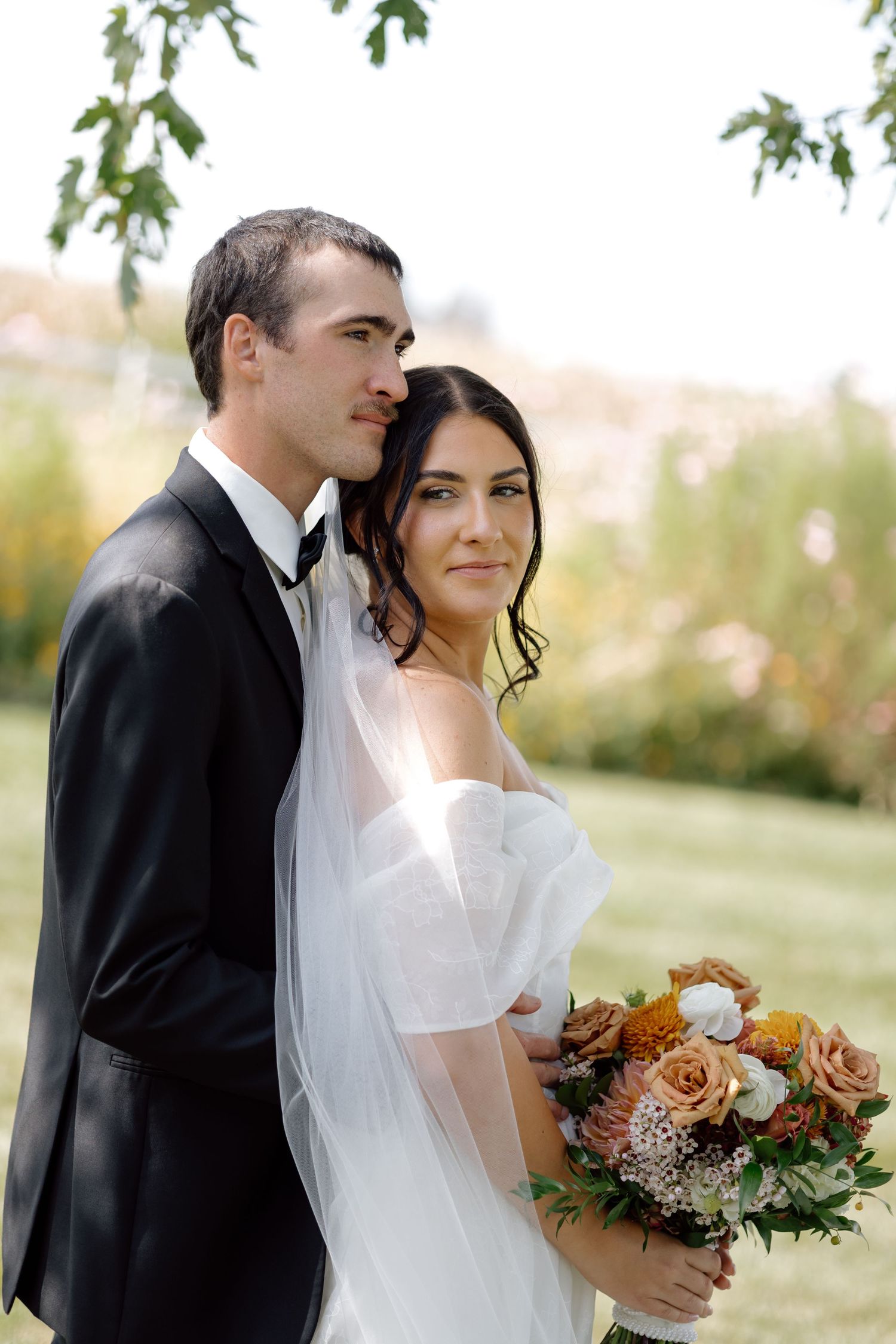 Elegant bride and groom posing together outdoors, bride holding autumn bouquet of roses and wildflowers.