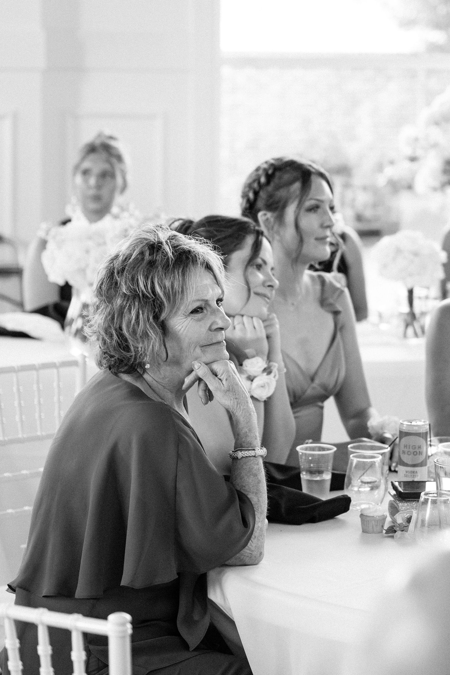 Black and white photo of wedding guests seated at a reception table, smiling and watching the ceremony.