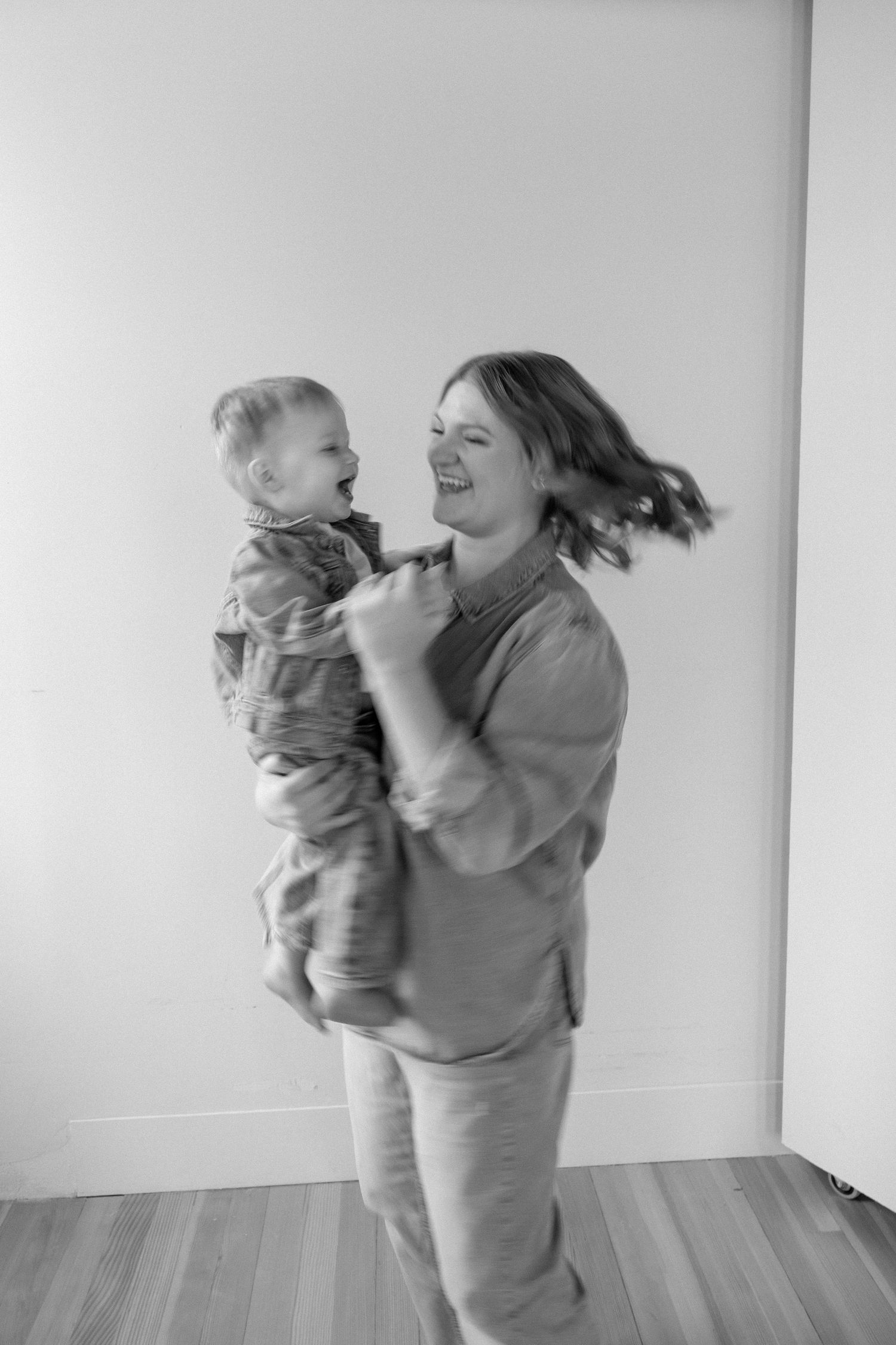 Black and white photo of laughing mother spinning happy toddler in bright white room.