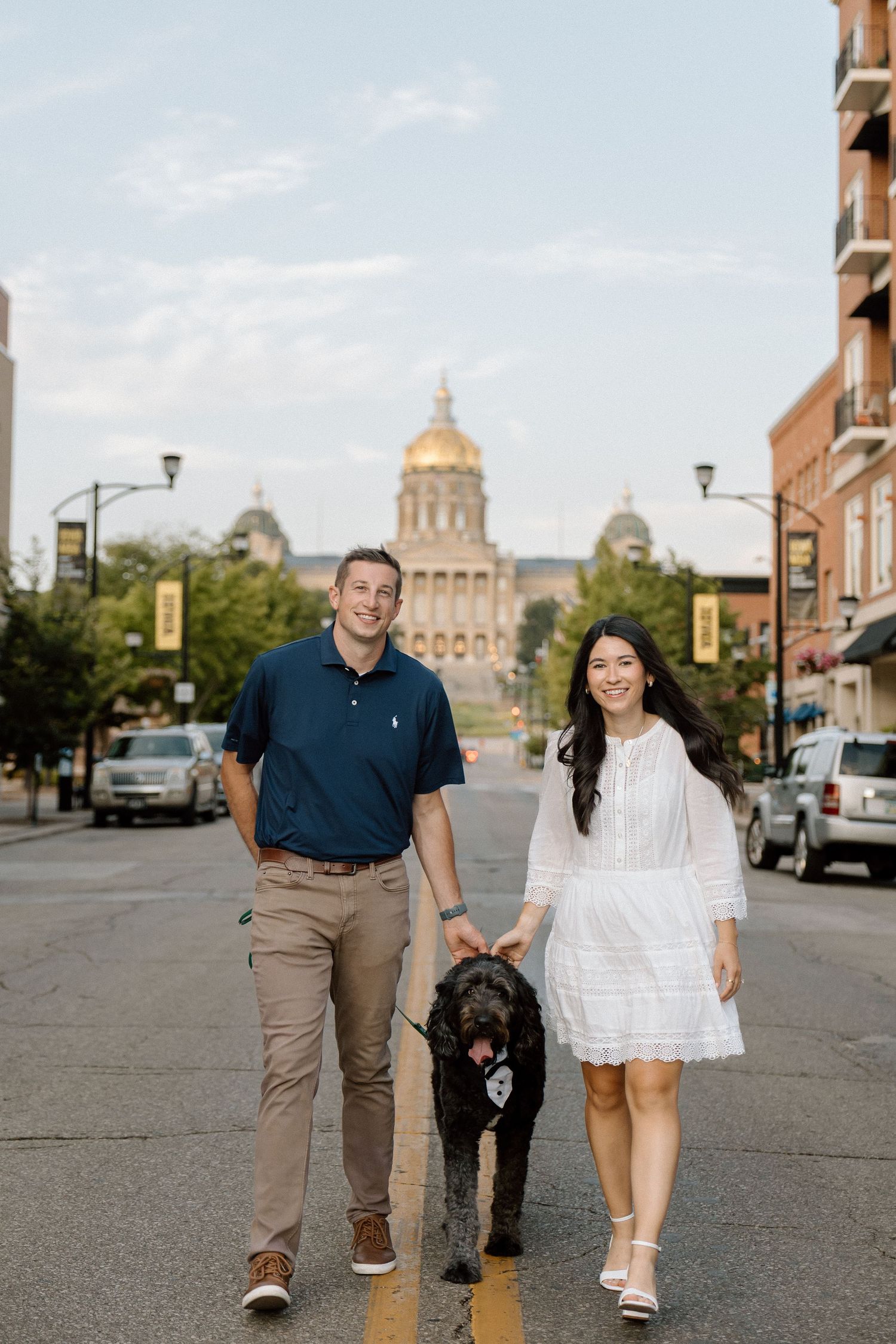 Smiling couple walking their black doodle dog on a street with the Iowa State Capitol building in the background.