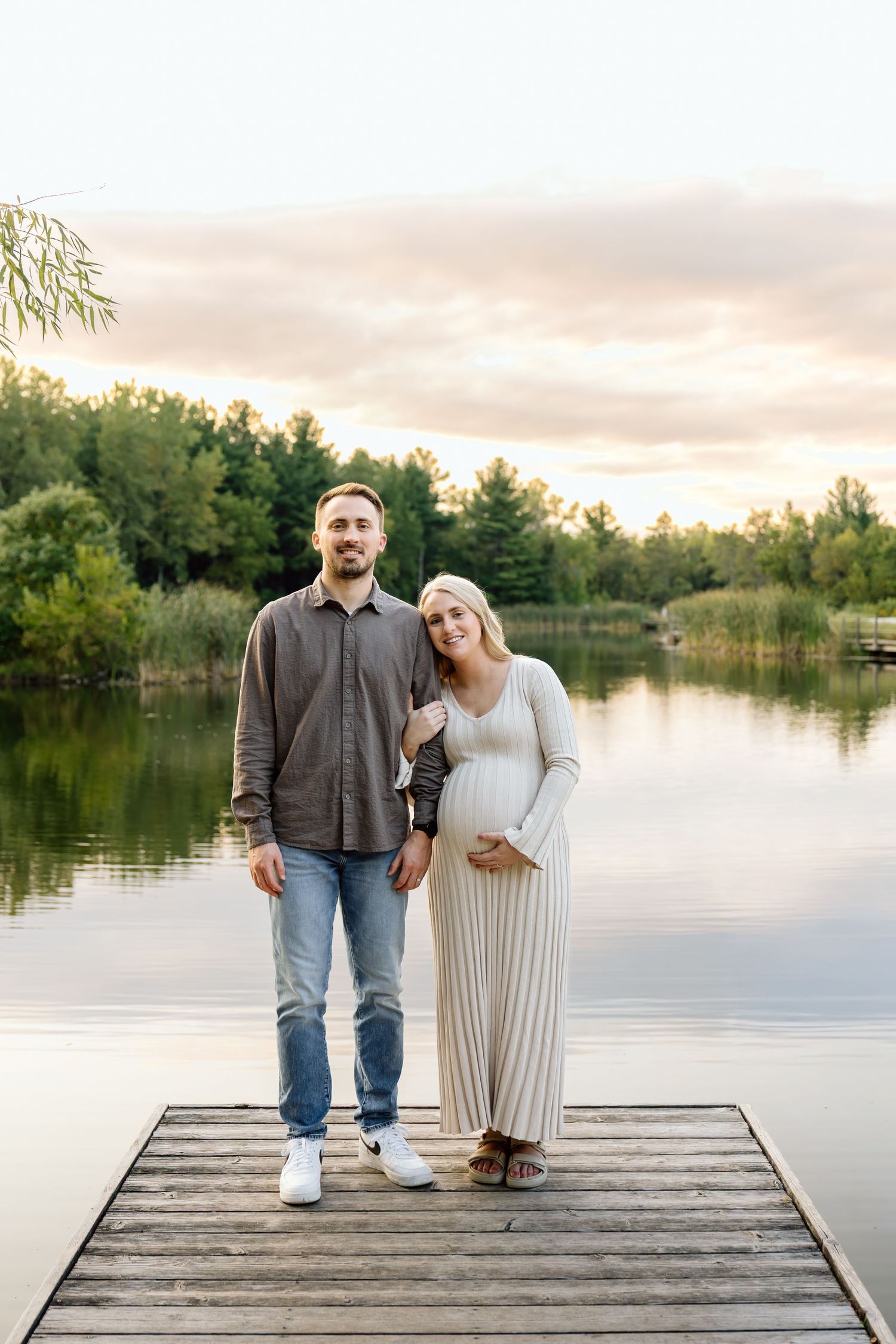 Pregnant couple standing on wooden dock at sunset with calm lake and trees behind them.