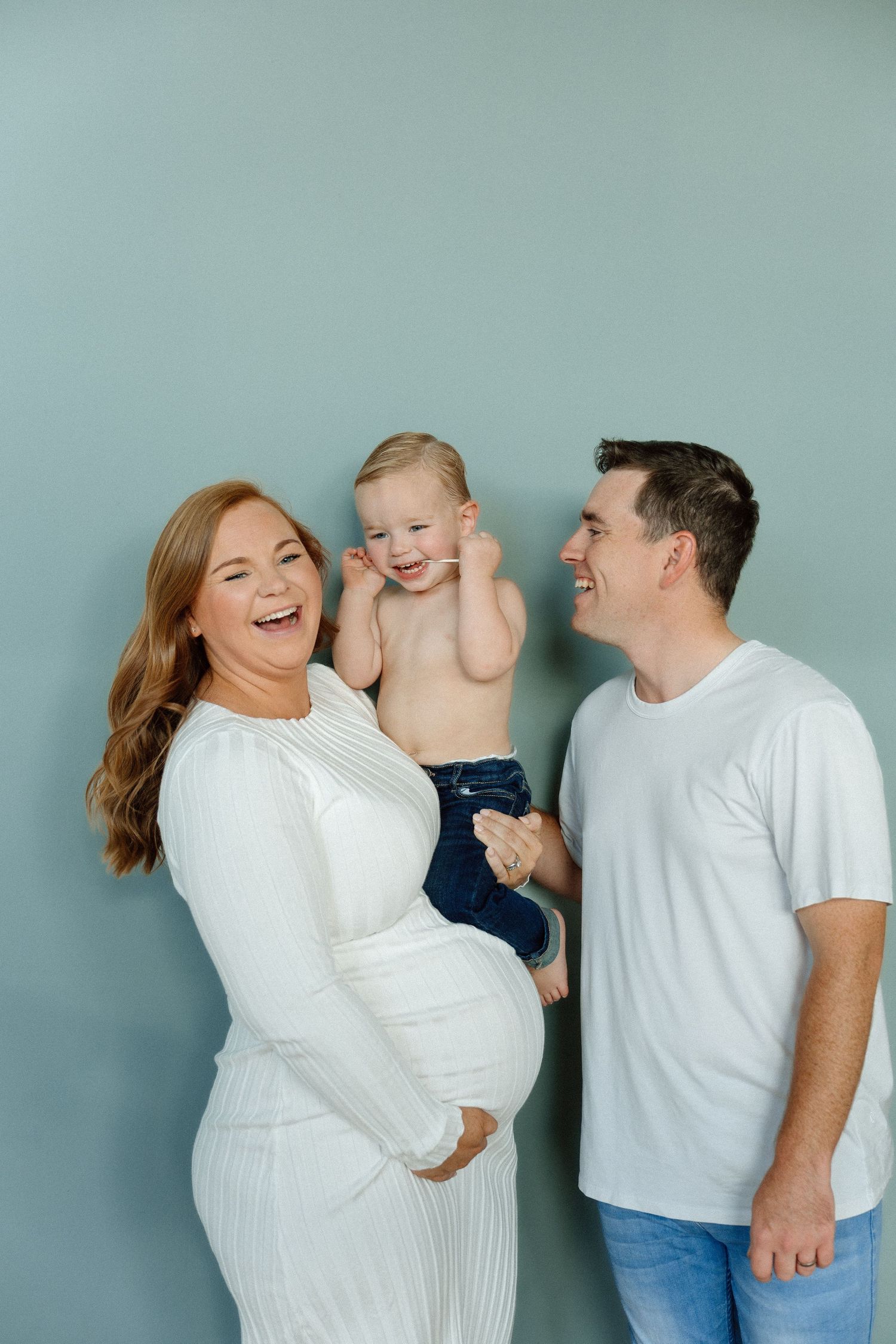 Joyful pregnant woman in white dress and husband laughing with their toddler against a blue wall.