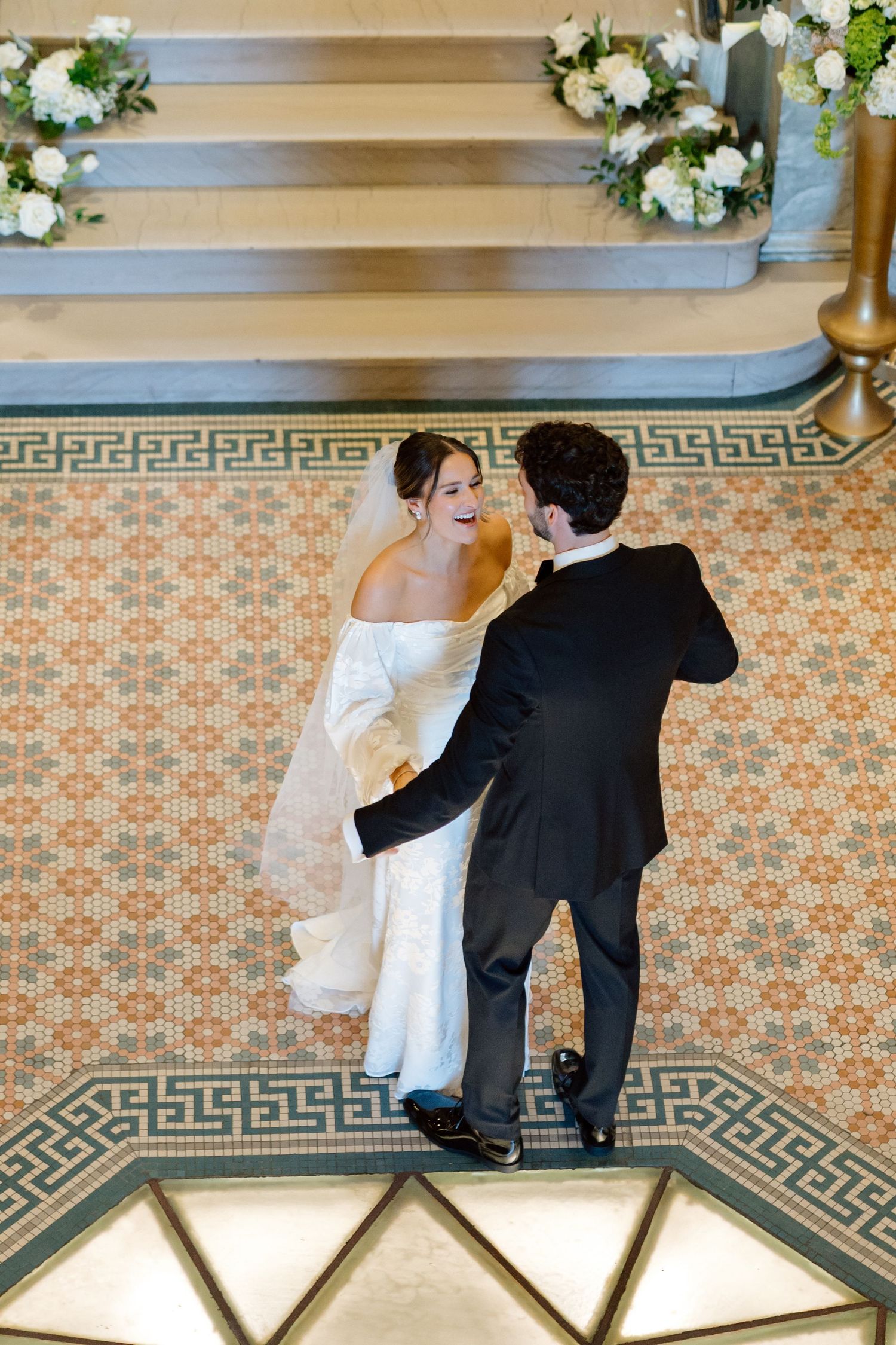Bride and groom share joyful first dance on mosaic tile floor with floral arrangements nearby.