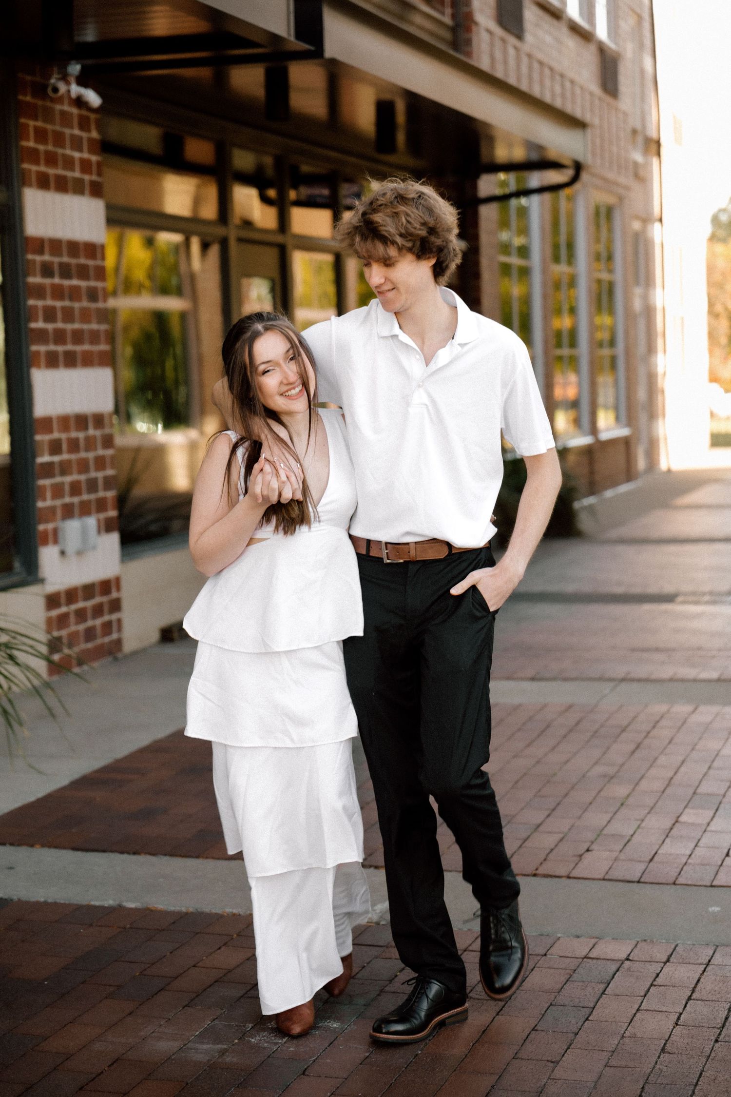 A young couple in coordinating white outfits pose together on a brick sidewalk outside a building.