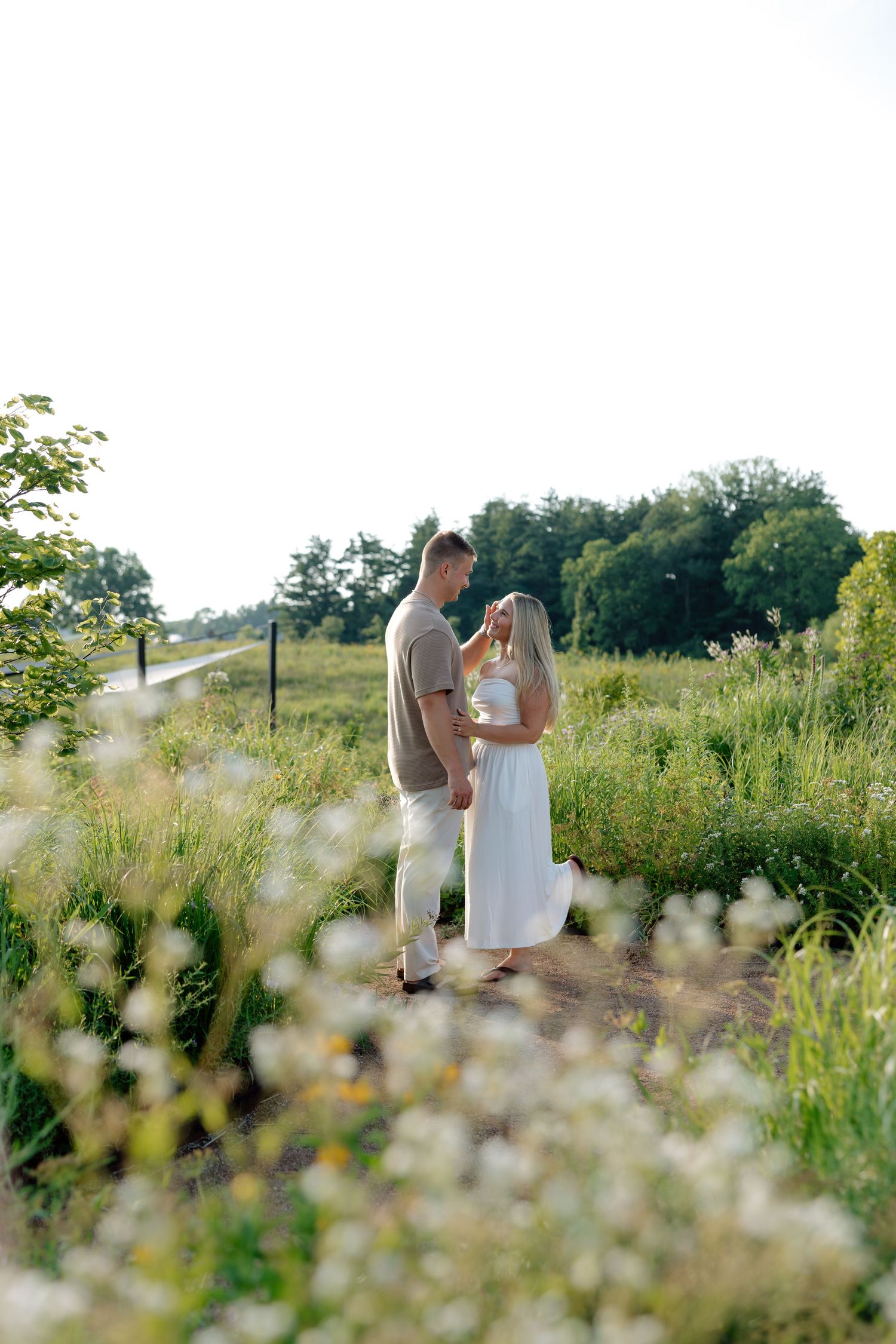 A couple embraces on a rural path surrounded by wildflowers and green trees on a bright summer day.