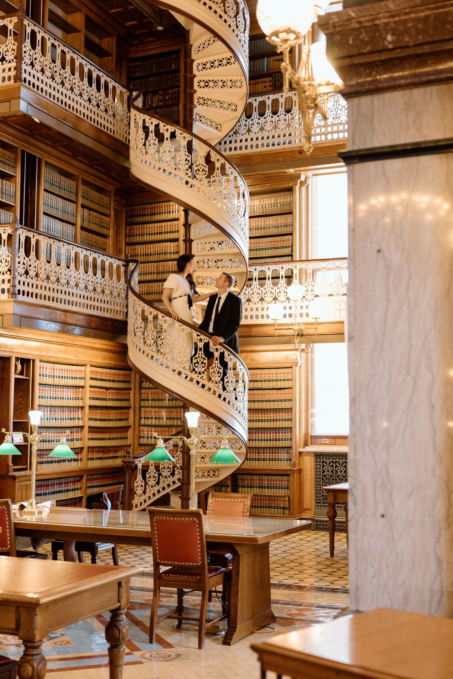 Couple posing on an ornate spiral staircase in a beautiful wood-paneled library with floor-to-ceiling bookshelves.