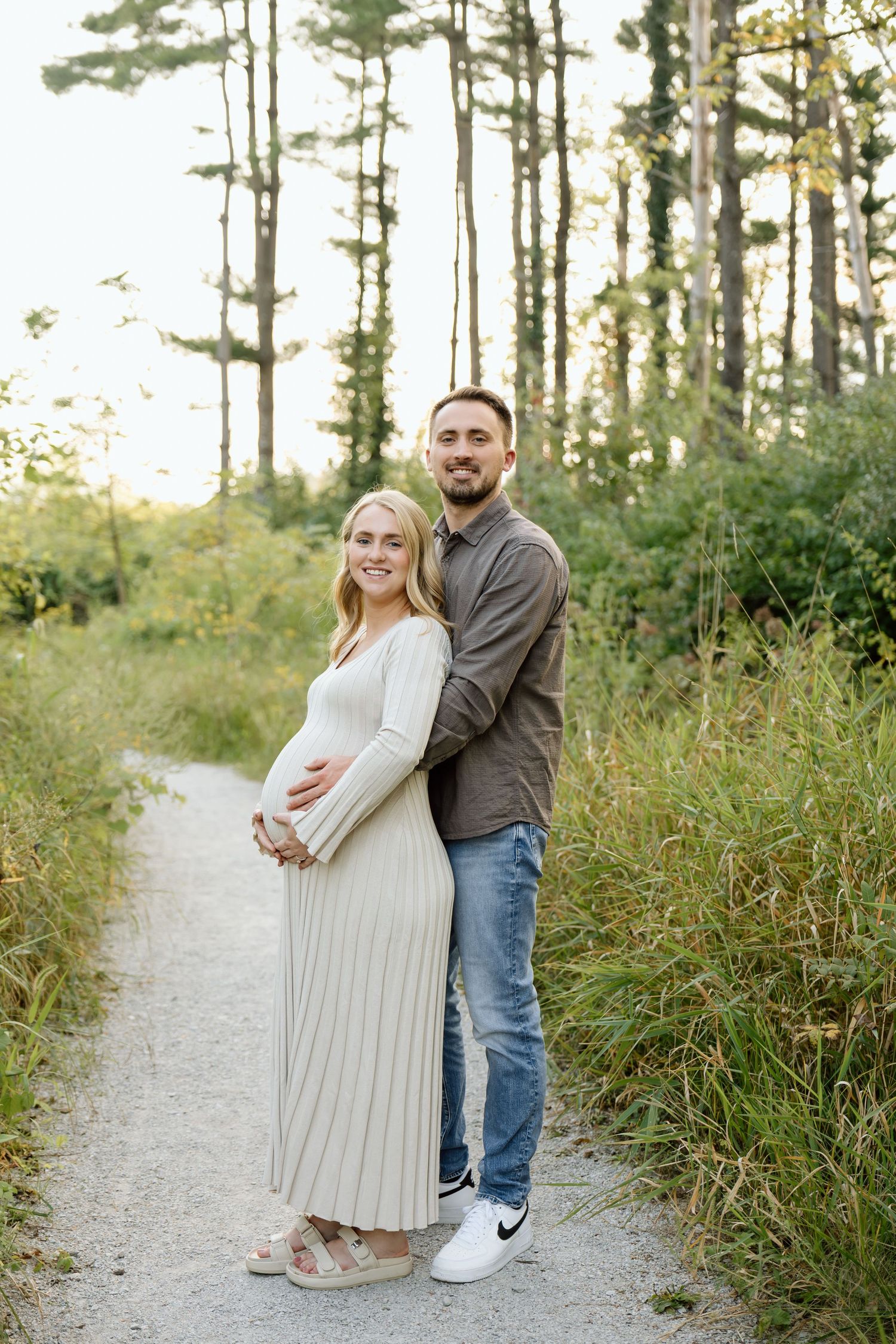 Expecting couple embracing on nature path at sunset, woman in cream ribbed maternity dress.