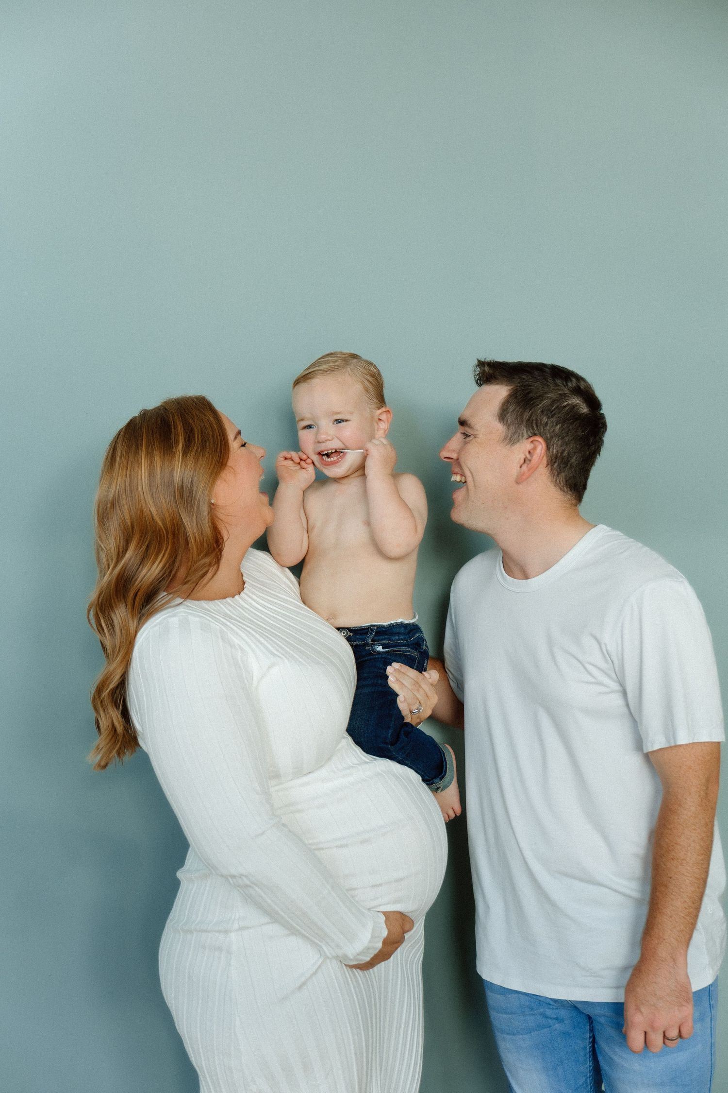 Smiling family with pregnant mother in white dress holding toddler beside father against blue wall.