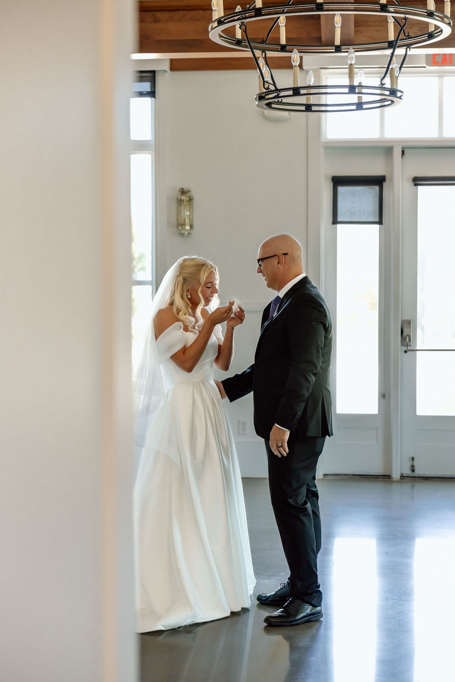 Bride in off-shoulder wedding gown shares tender first look moment with bald groom indoors.