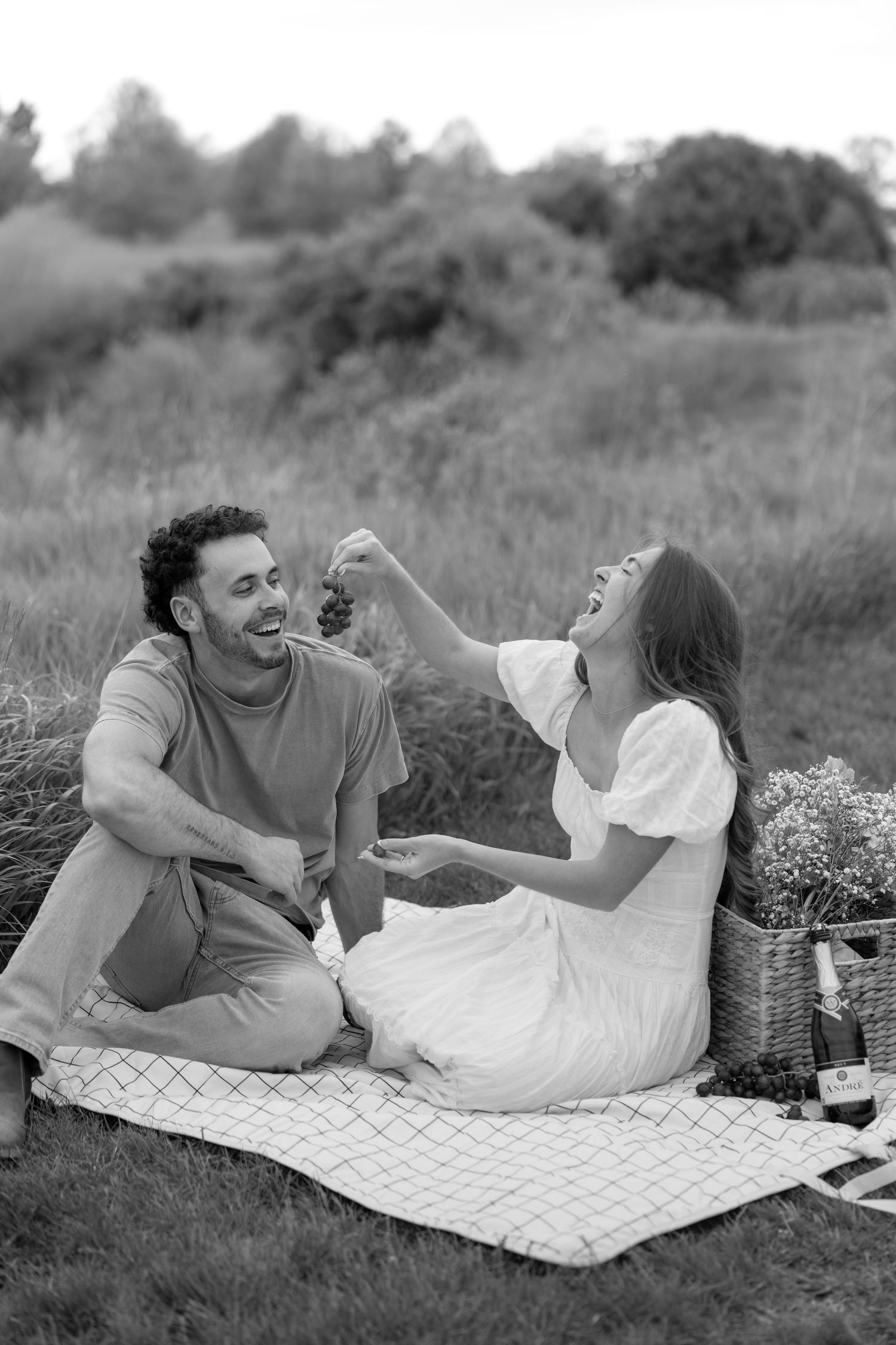 Black and white photo of a couple enjoying a romantic outdoor picnic, woman laughing while feeding man grapes on a blanket.