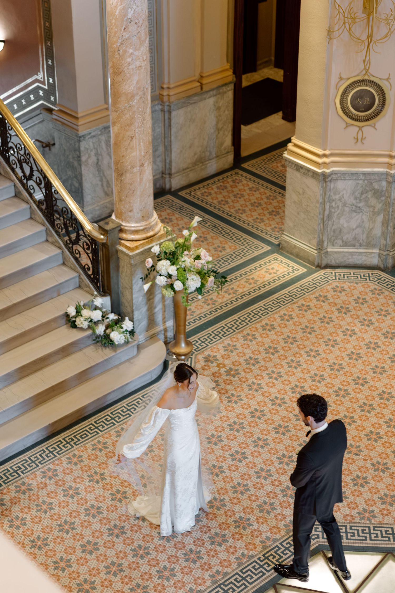 Aerial view of bride and groom sharing first look near grand staircase with floral arrangement.