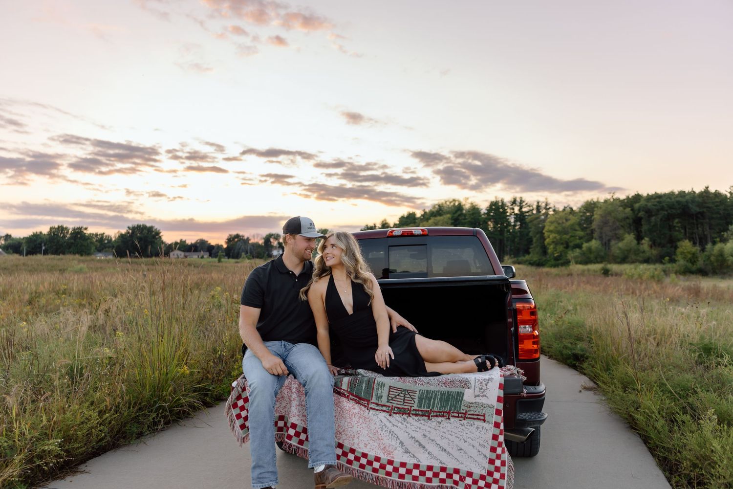 Couple sitting on truck tailgate with checkered blanket at sunset in a scenic field.
