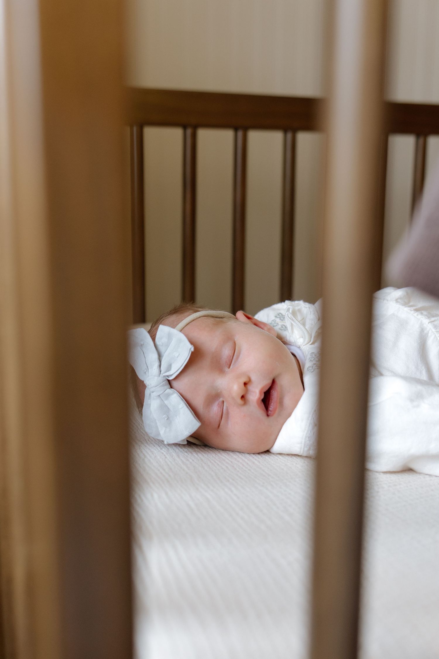 Sleeping newborn baby with a white bow headband in a wooden crib.