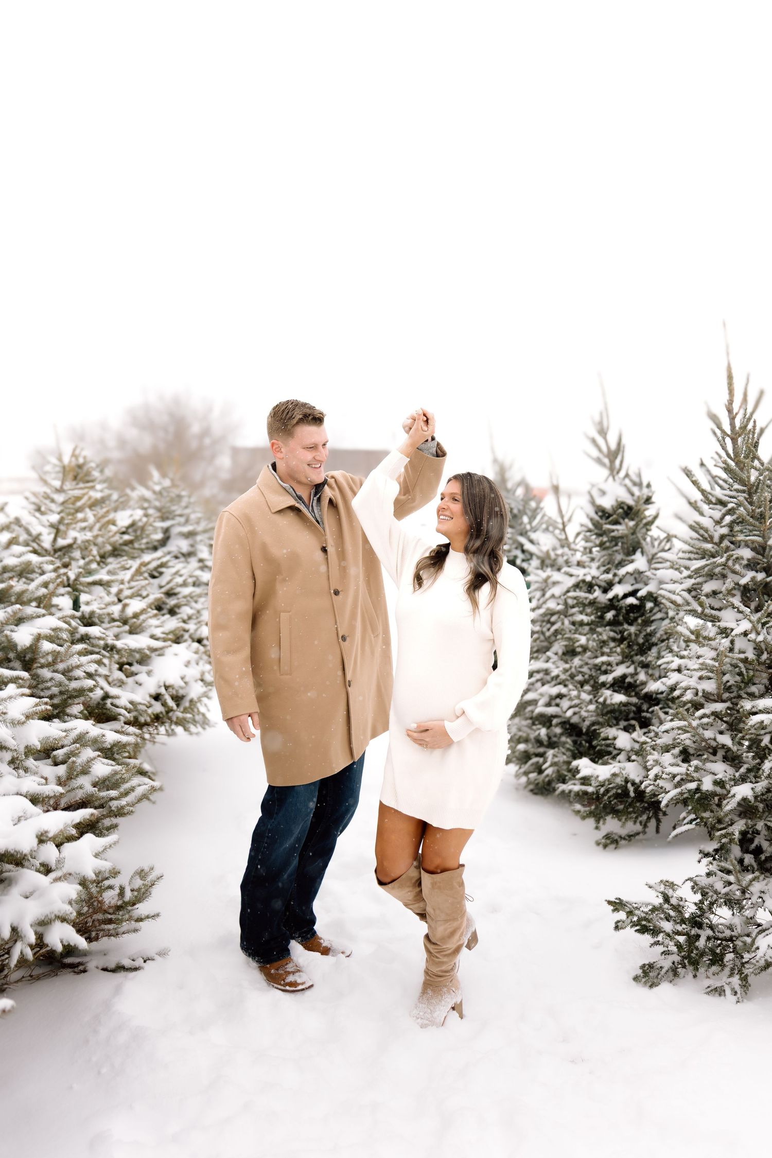 Couple dancing in snowy Christmas tree farm, pregnant woman in white dress and tall boots.