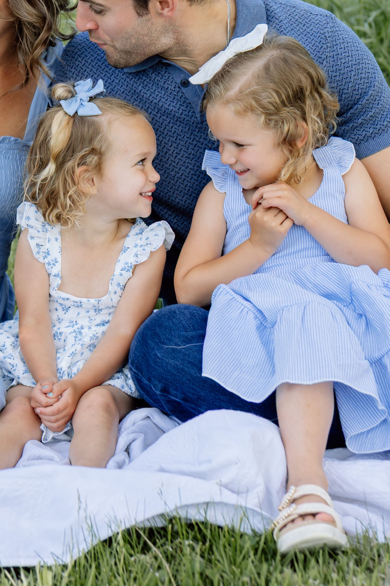 Two young curly-haired sisters in blue dresses smiling at each other during an outdoor family photo.