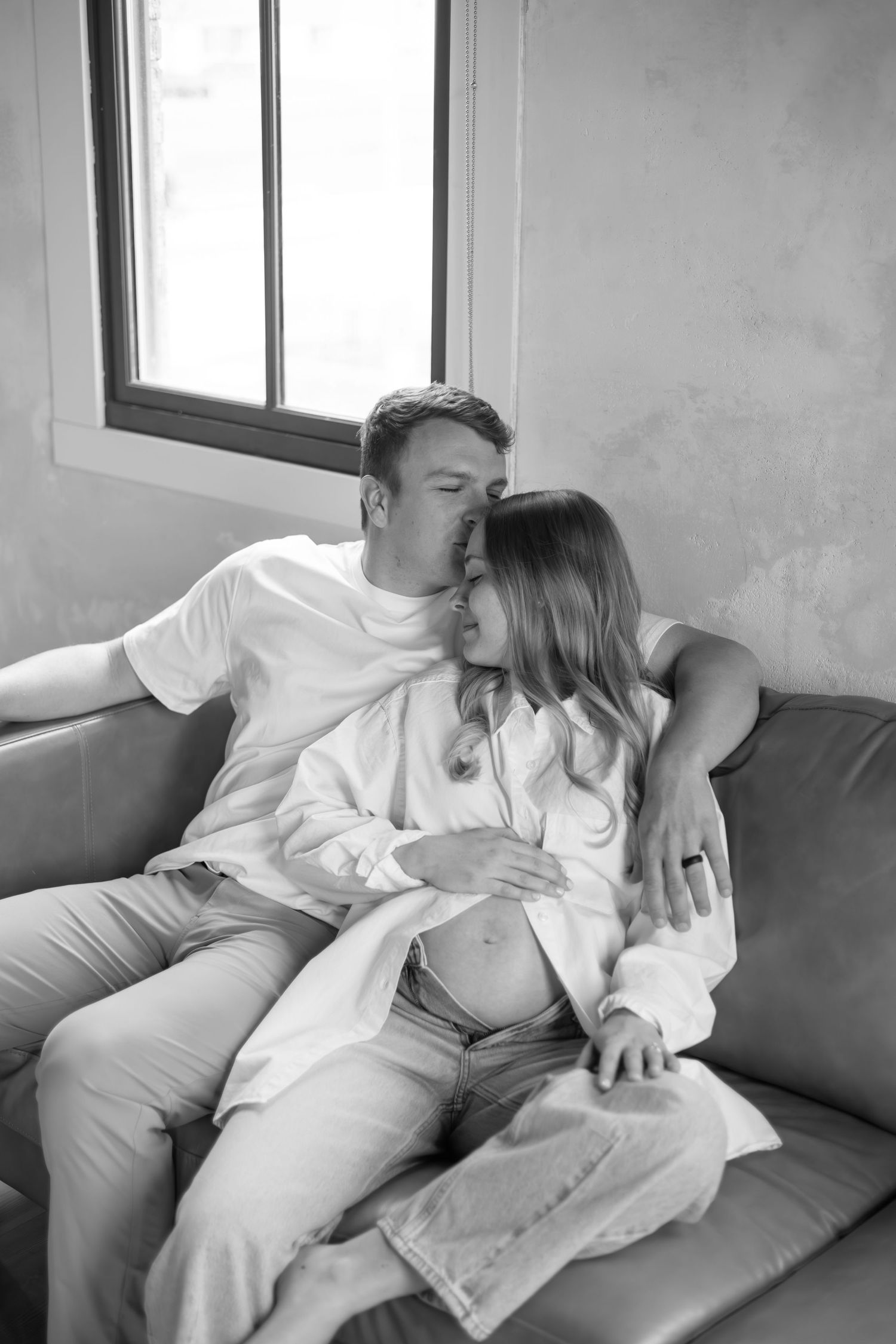 Black and white photo of a couple sitting on a couch, man kissing woman's head as she shows her bump.