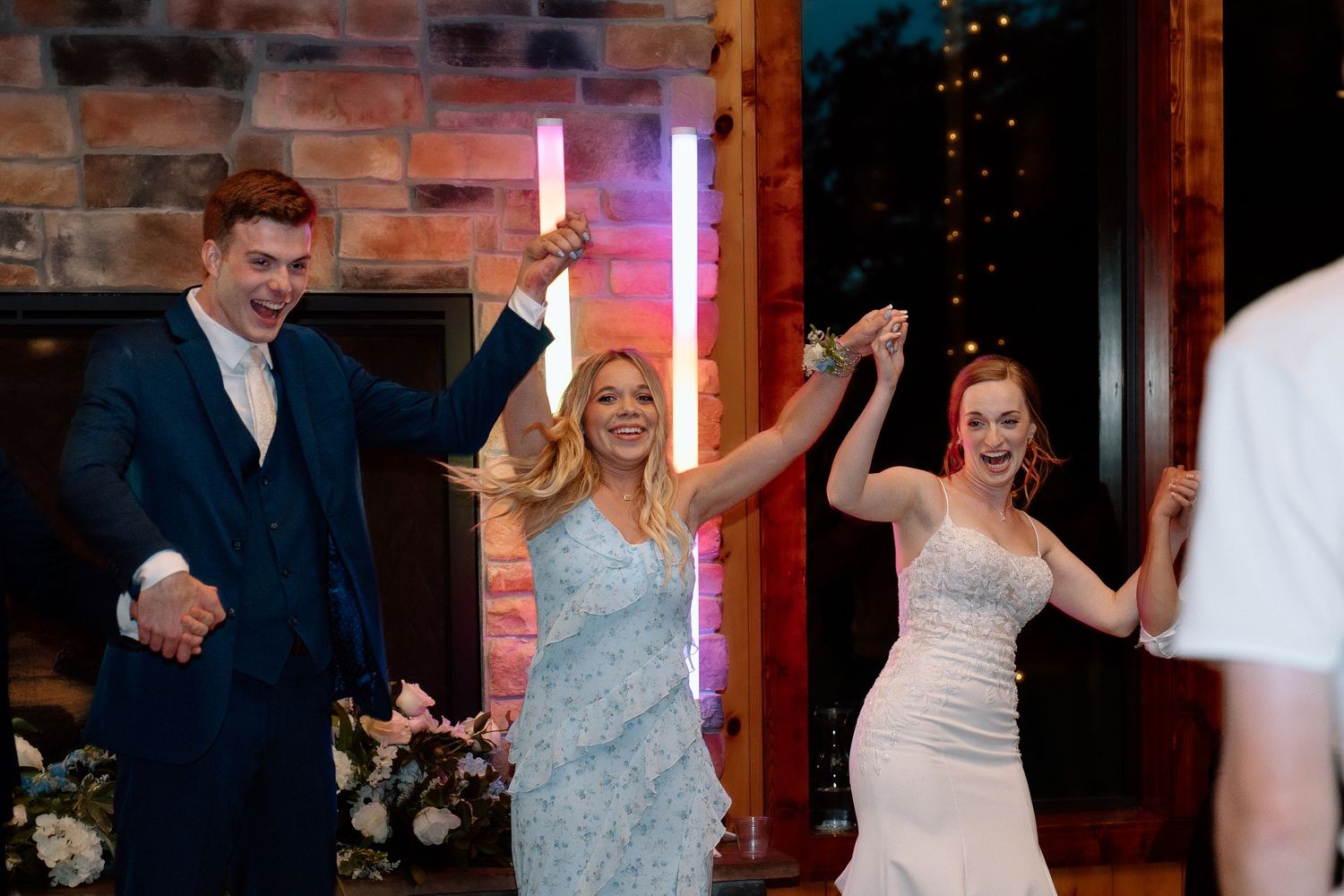 A bride and guests celebrate with light-up wands near a stone fireplace at a wedding reception.