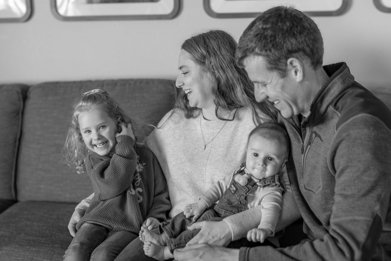 Black and white family photo of parents smiling with toddler girl and baby boy sitting on a cozy couch.