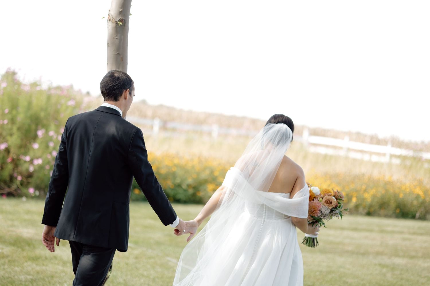 Bride and groom walk hand in hand through a garden, bride carrying a colorful autumn bouquet.