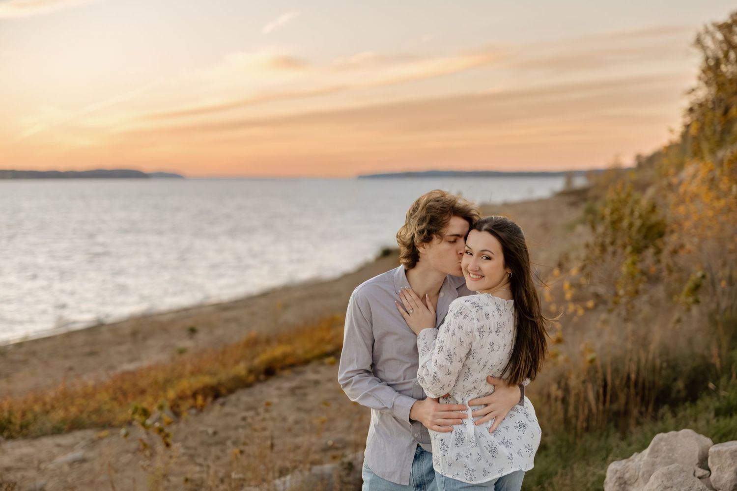 Couple embracing at sunset by the water, golden hour light, romantic outdoor engagement photo on rocky shoreline.