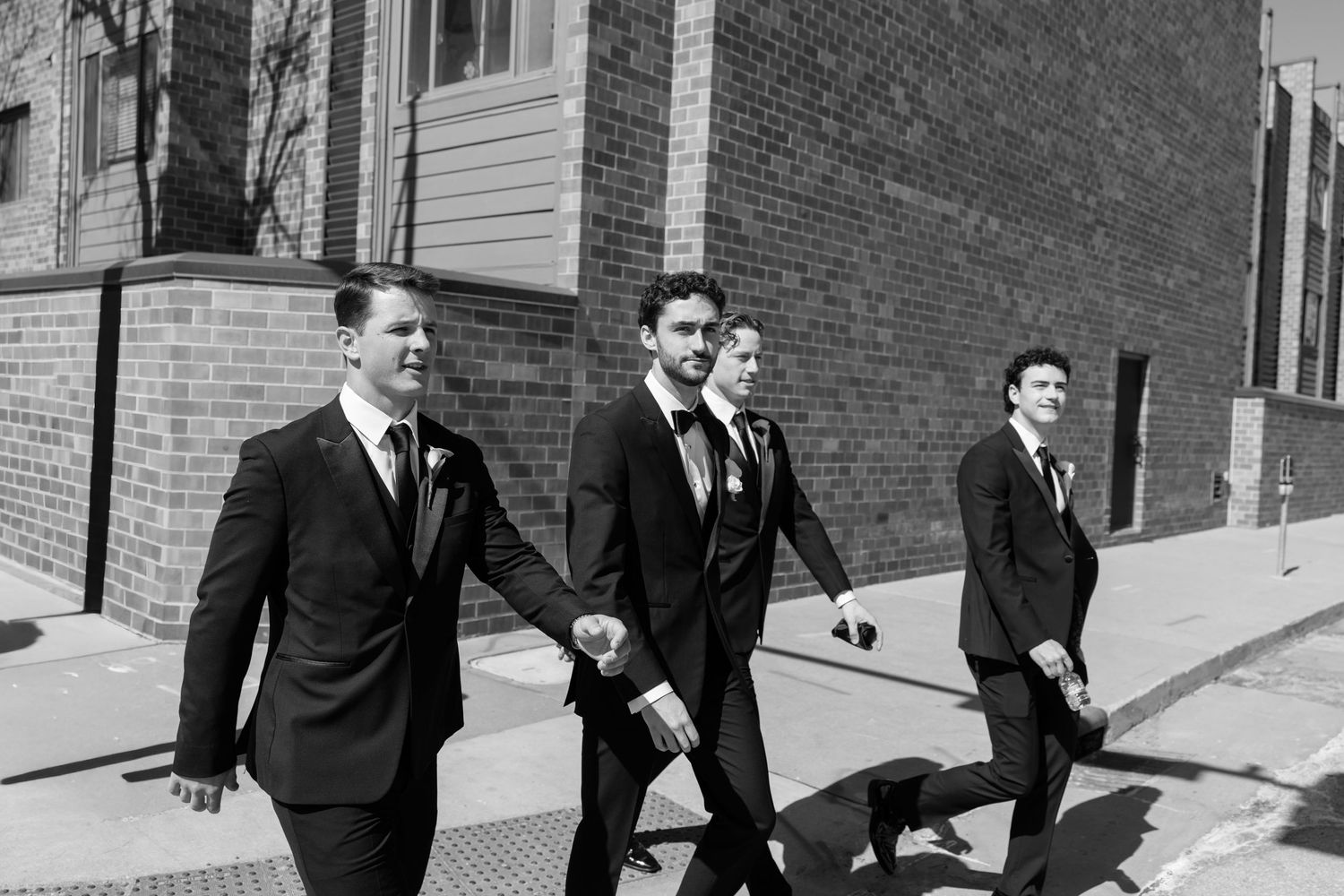 Three groomsmen in black suits walking confidently down a sunny urban street on a wedding day.
