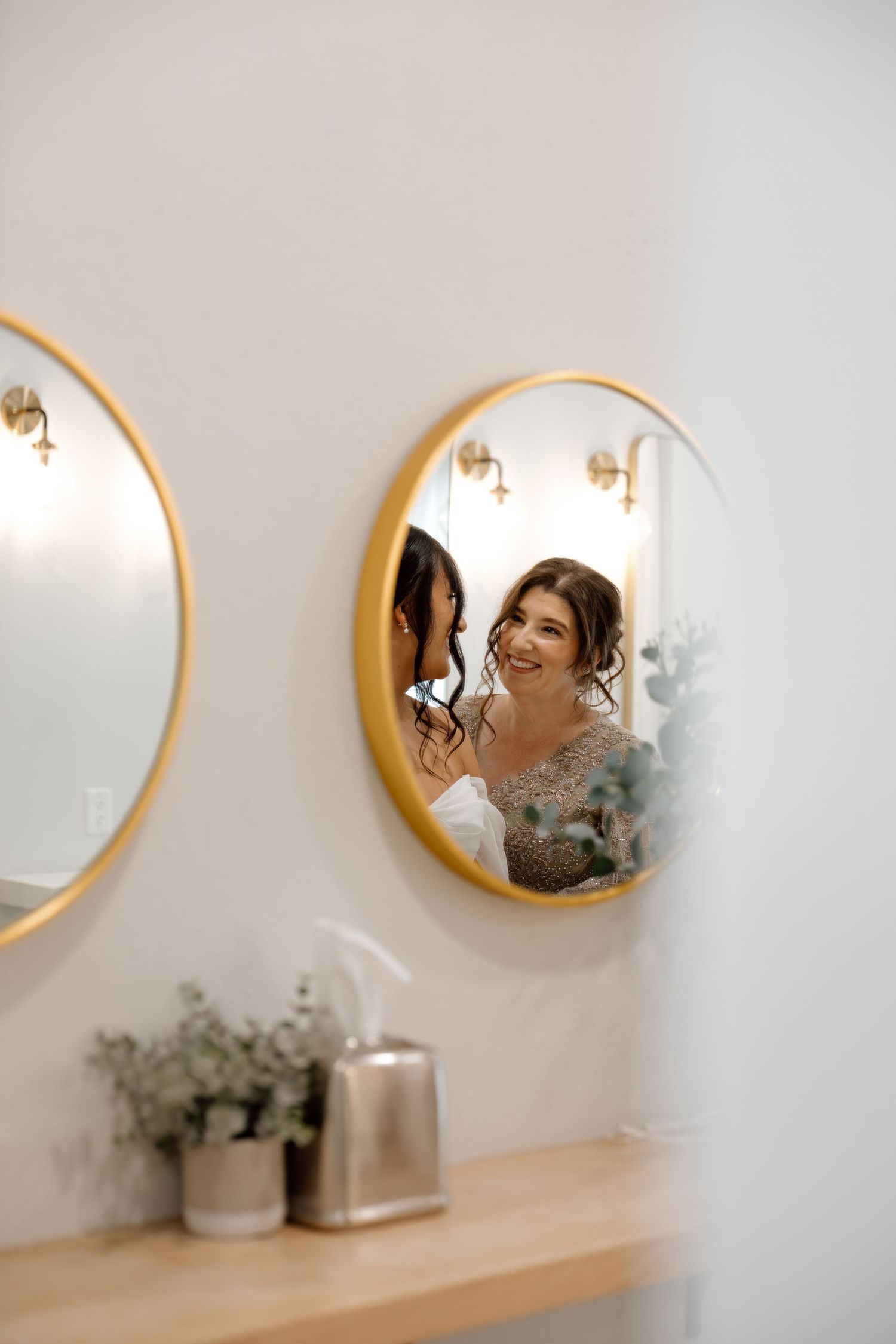 Bride smiling in gold-framed mirror reflection while bridesmaid helps with hair in bright white bridal suite.
