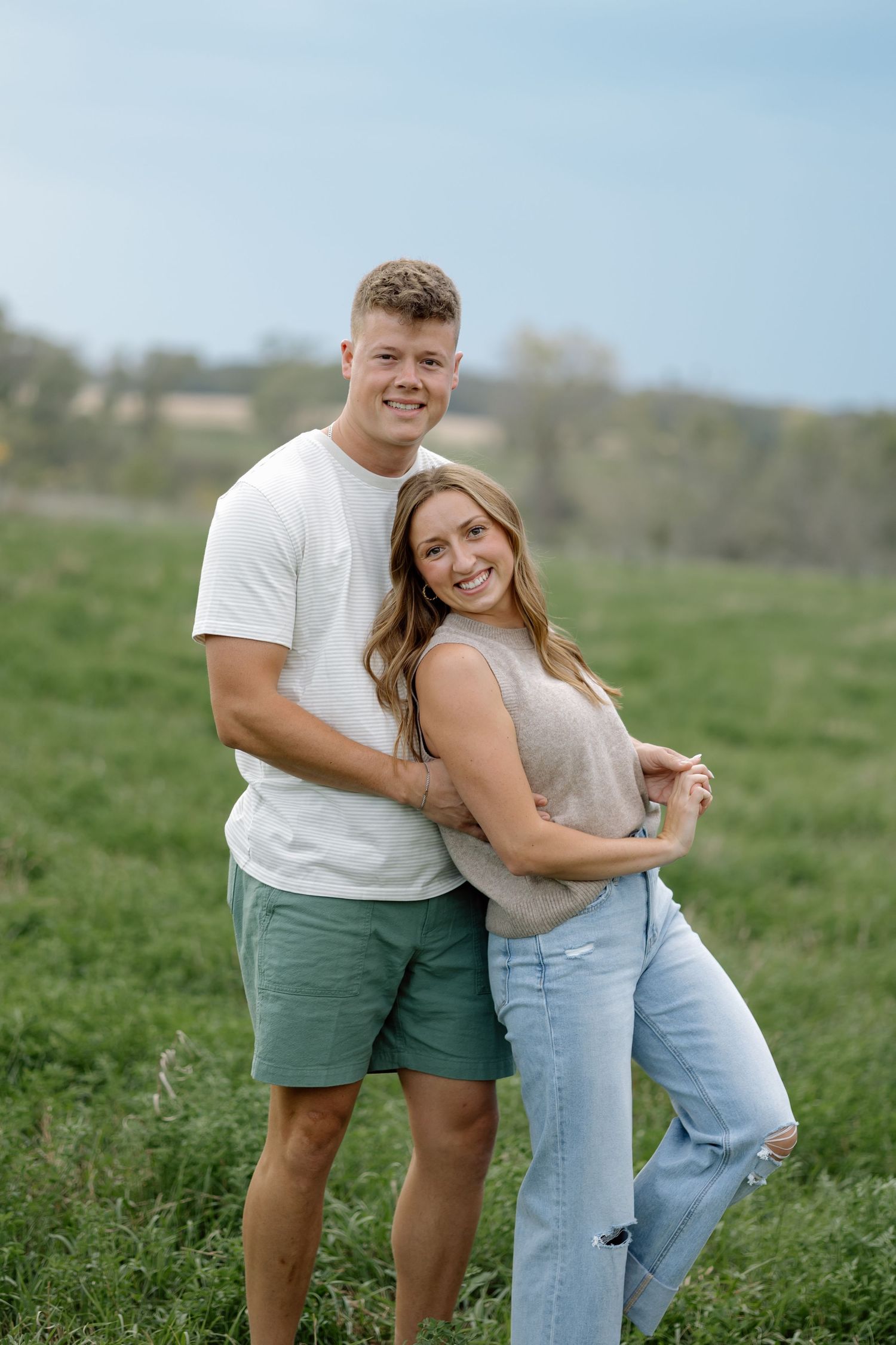 Smiling couple embracing in green meadow, man in white tee and green shorts, woman in jeans.