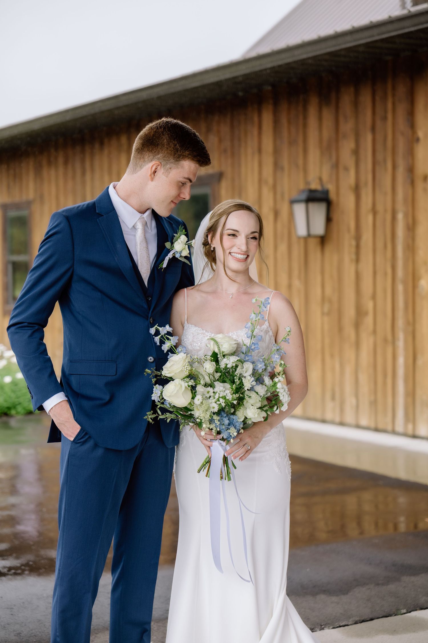 Groom in navy suit and bride in white gown holding a blue and white bouquet pose outside a rustic venue.