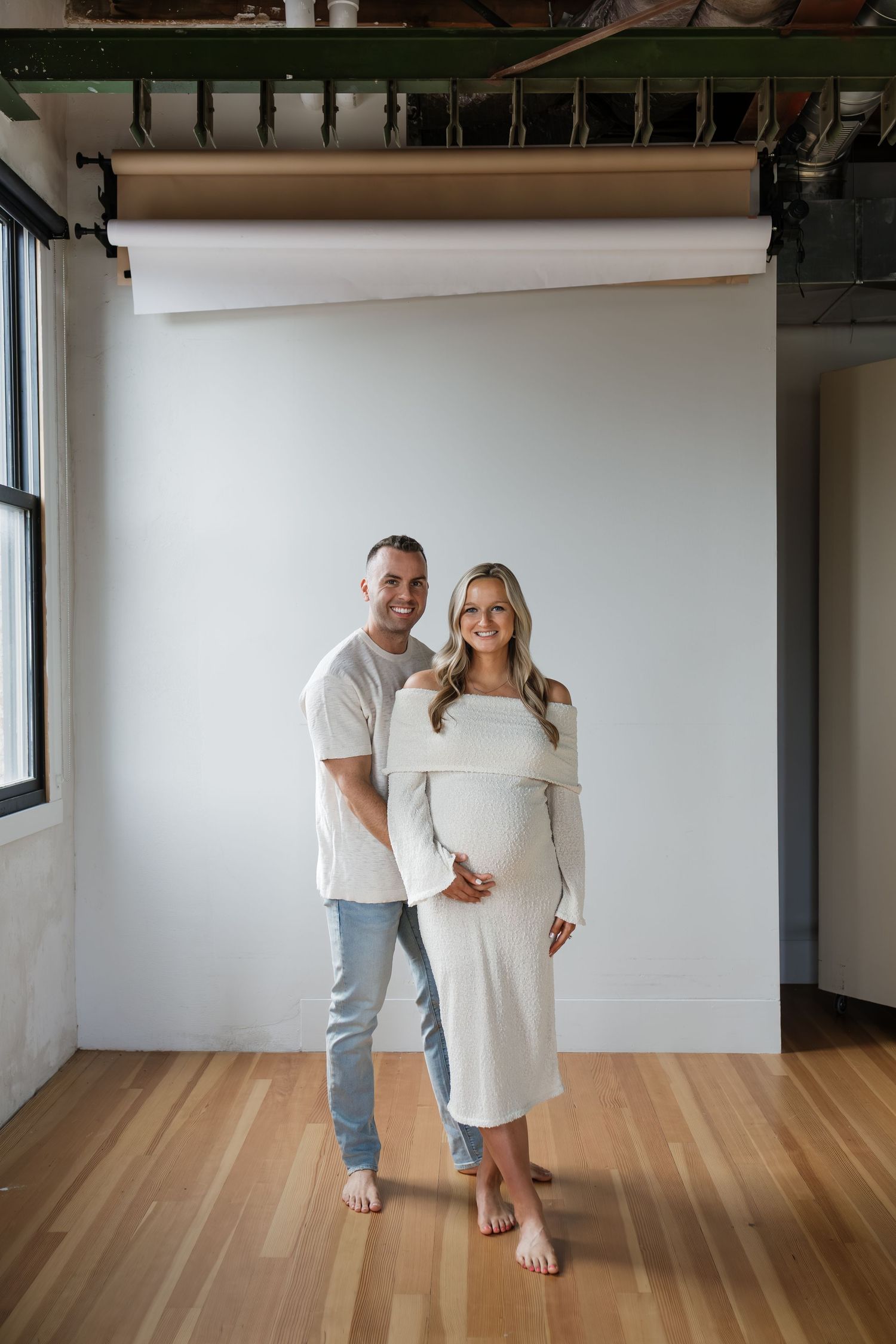 Smiling couple posing for maternity photo, woman in cream dress cradling pregnant belly.