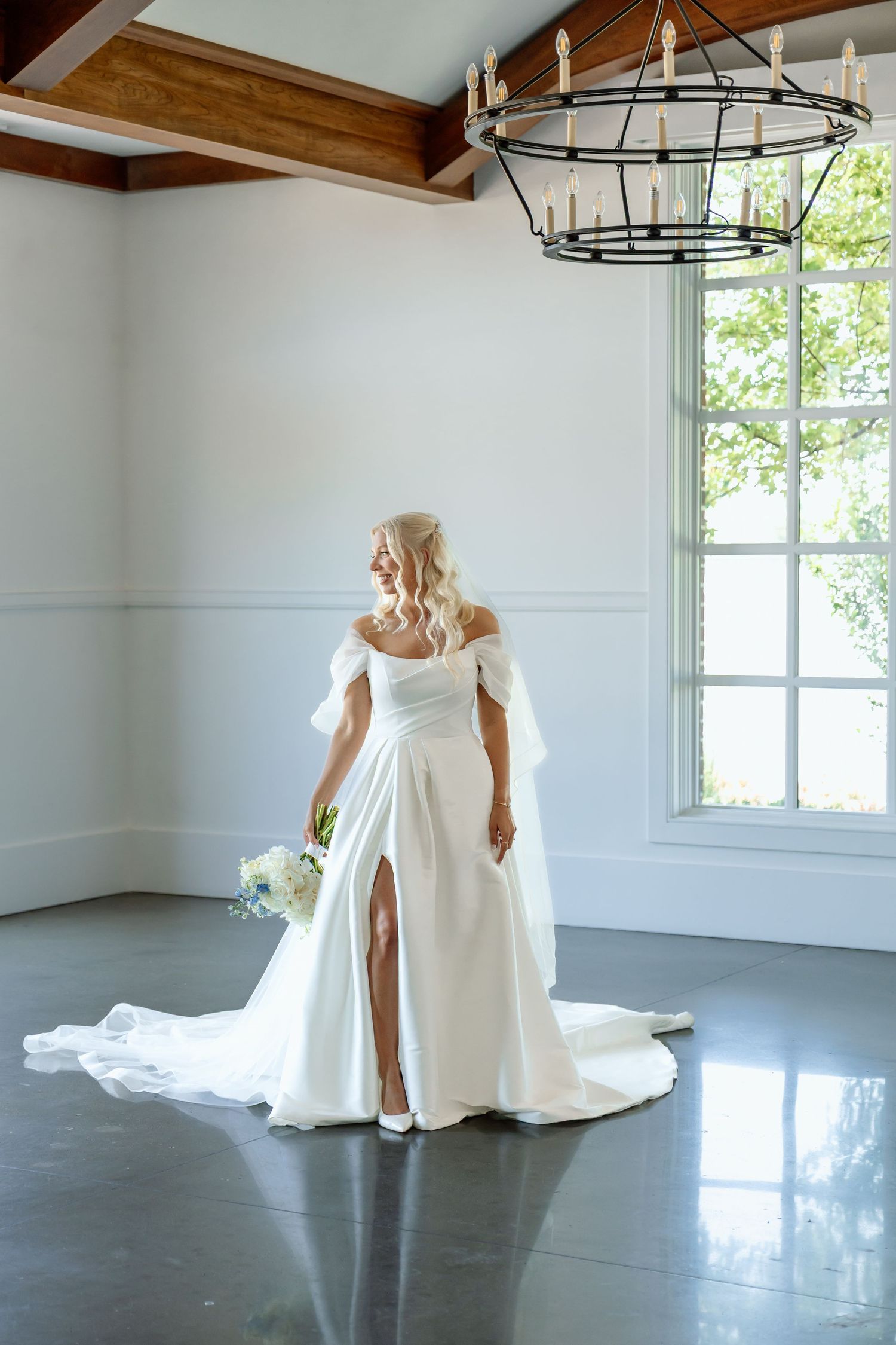 Elegant bride in off-shoulder white gown with slit holding bouquet under a chandelier at The Monroe wedding venue in West Des Moines, Iowa.