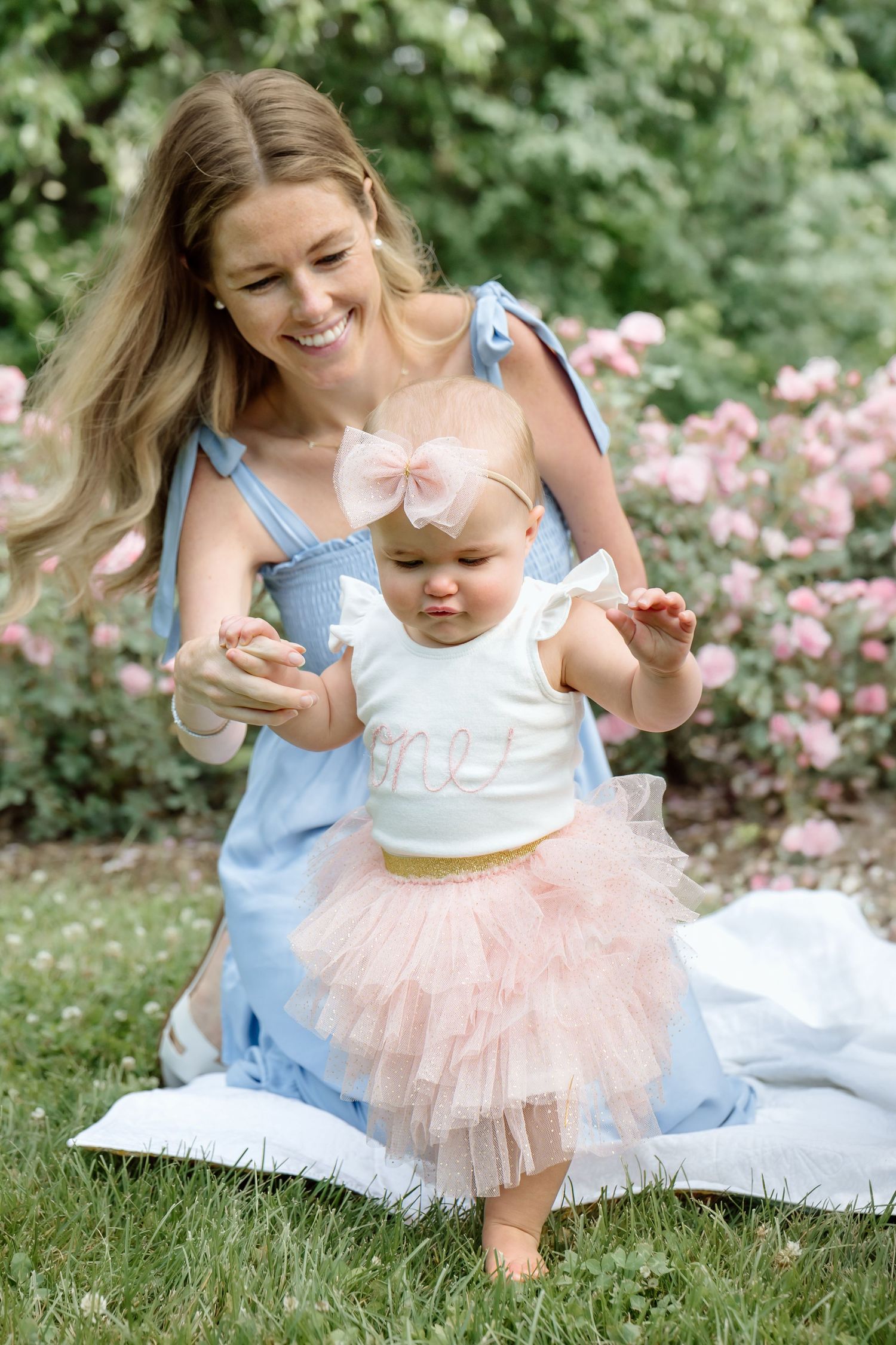 Smiling blonde mother in blue dress helps her baby girl in pink tutu stand on a garden blanket.