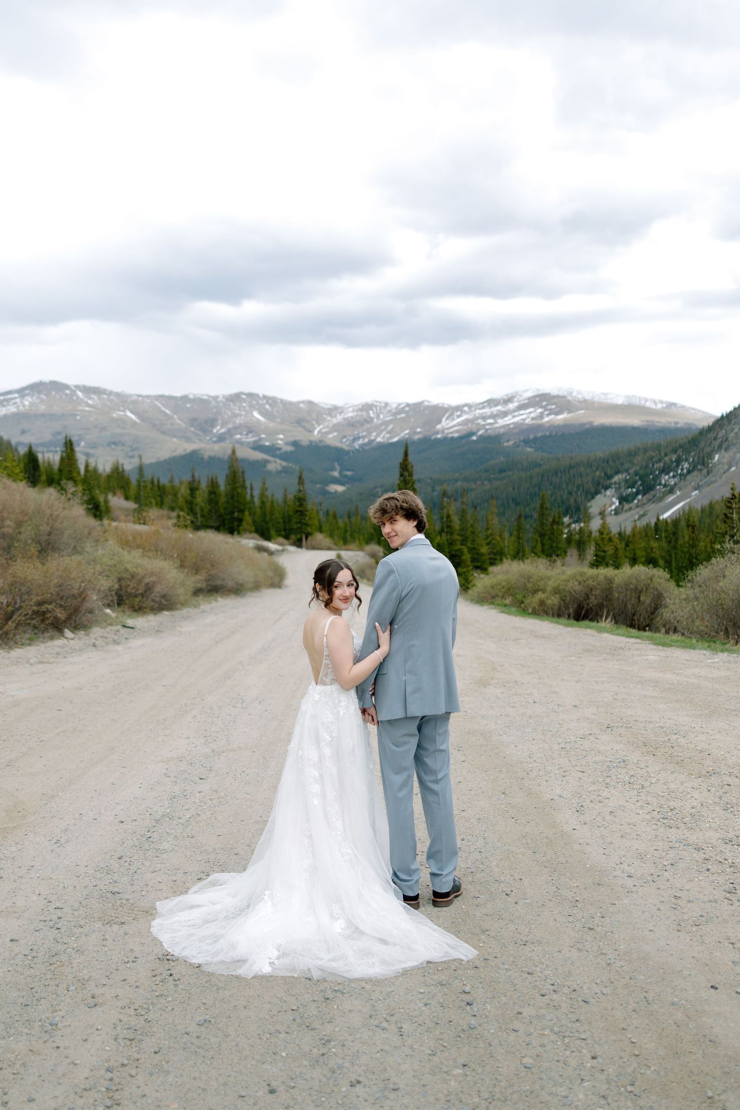 Bride and groom holding hands on a mountain dirt road with snow-capped peaks and evergreen trees in Colorado.