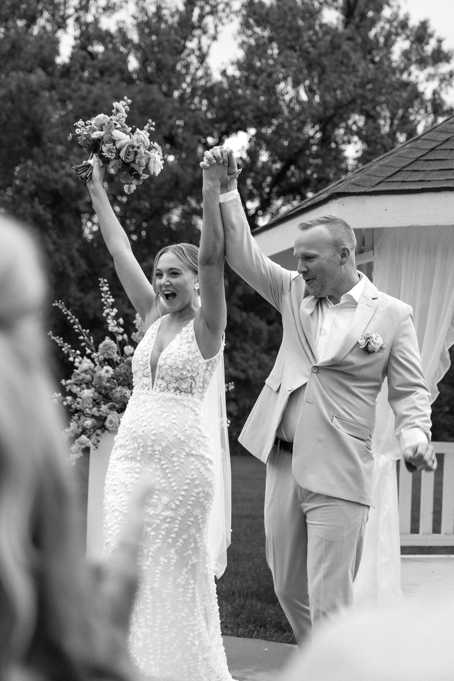 Black and white photo of joyful newlyweds raising hands after outdoor wedding ceremony.